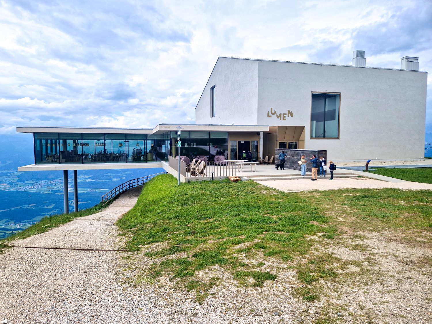 Things To Do In The Dolomites In Winter: Ski And No Ski 16 A modern white building labeled "LUMEN" sits on a grassy hilltop, with large glass windows overlooking a scenic mountain landscape. Several people stand outside under a cloudy sky.