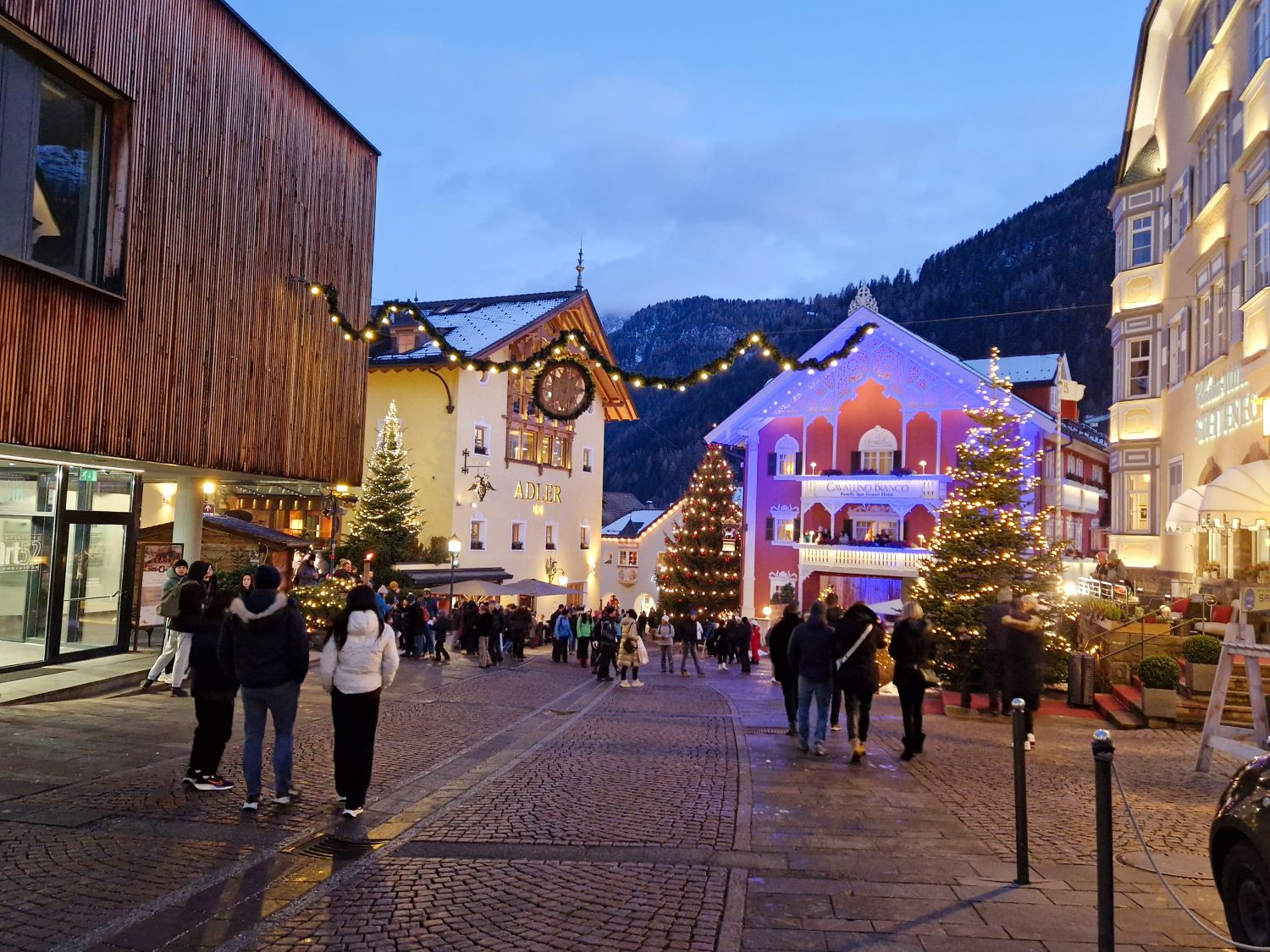 Dolomites in Winter: Weather, Skiing, Hiking, and more 20 Festive town square at dusk with people walking along a cobblestone street in Ortisei, Val Gardena lined with lit Christmas trees and glowing string lights overhead. Hotel buildings decorated with warm lights include one with a clock and the word "ADLER" and another reading "CAVALINO BANCO" set against dark mountain silhouettes in the background.