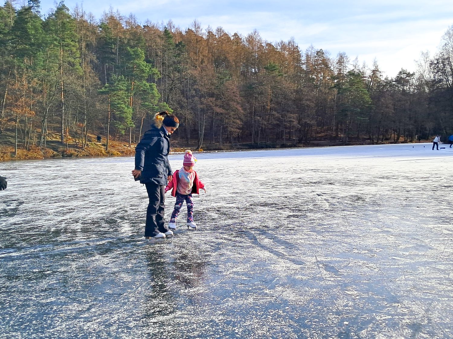 Things To Do In The Dolomites In Winter: Ski And No Ski 18 An adult and a child, both bundled up in winter clothes, hold hands while ice skating on a frozen lake surrounded by trees under a clear sky.