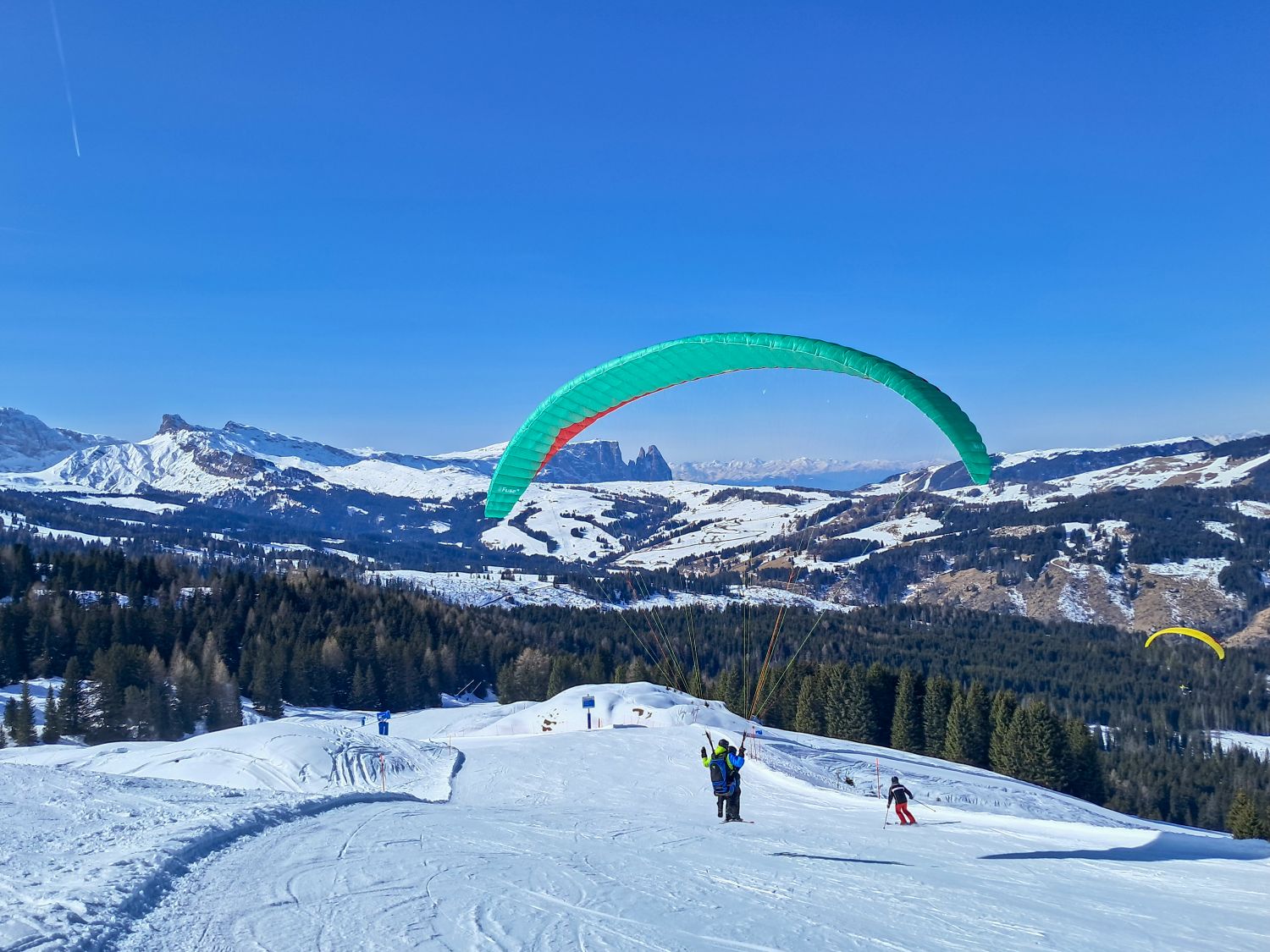 Dolomites in Winter: Weather, Skiing, Hiking, and more 16 a green paraglider prepared to launch on a snowy ridge in Monte Pana with wide alpine views of rolling hills and jagged peaks in the distance. Another skier moves downhill nearby while a second paraglider floats over the forested valley under a clear blue sky.