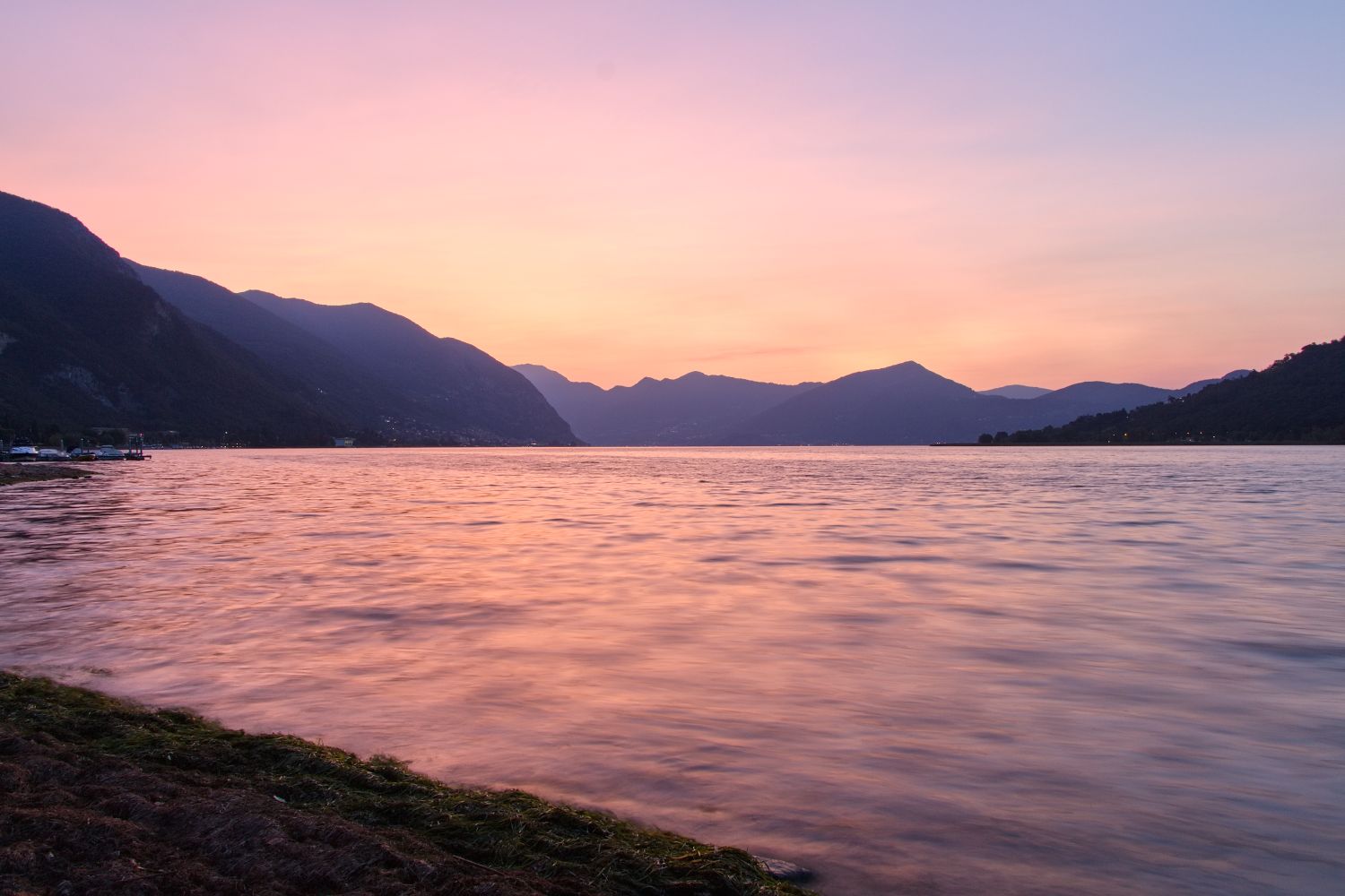 11 beautiful lakes in Northern Italy 2 A calm lake at sunrise with gentle waves, distant mountains silhouetted against a pink and orange sky, and a grassy shoreline in the foreground.