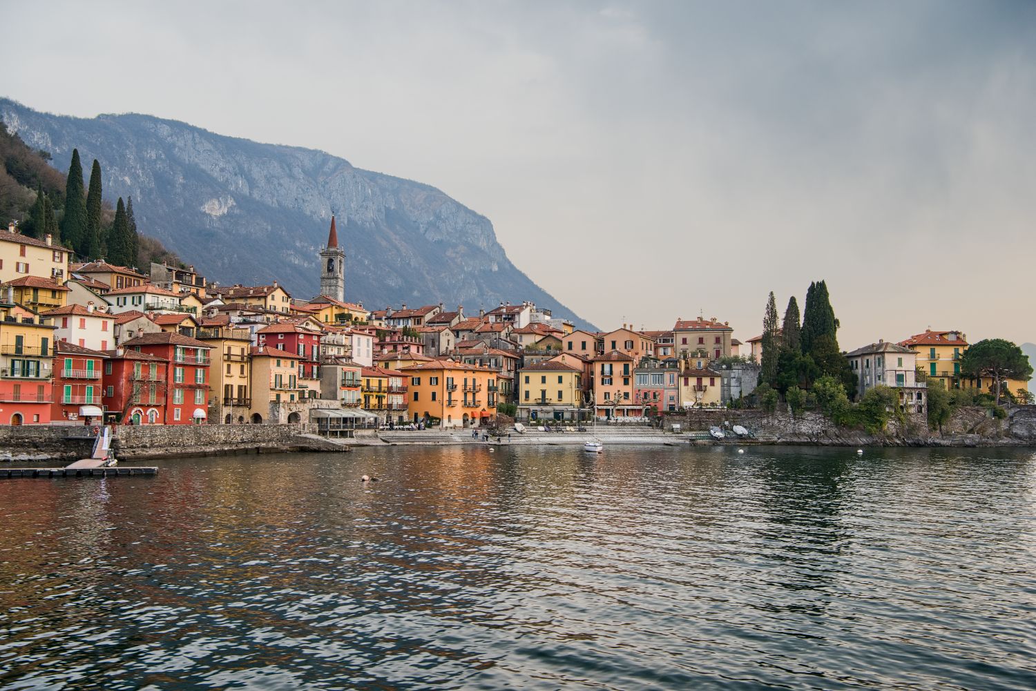11 beautiful lakes in Northern Italy 4 A picturesque lakeside village with colorful buildings, a church tower, and cypress trees, set against mountains and reflected in calm water under a cloudy sky.