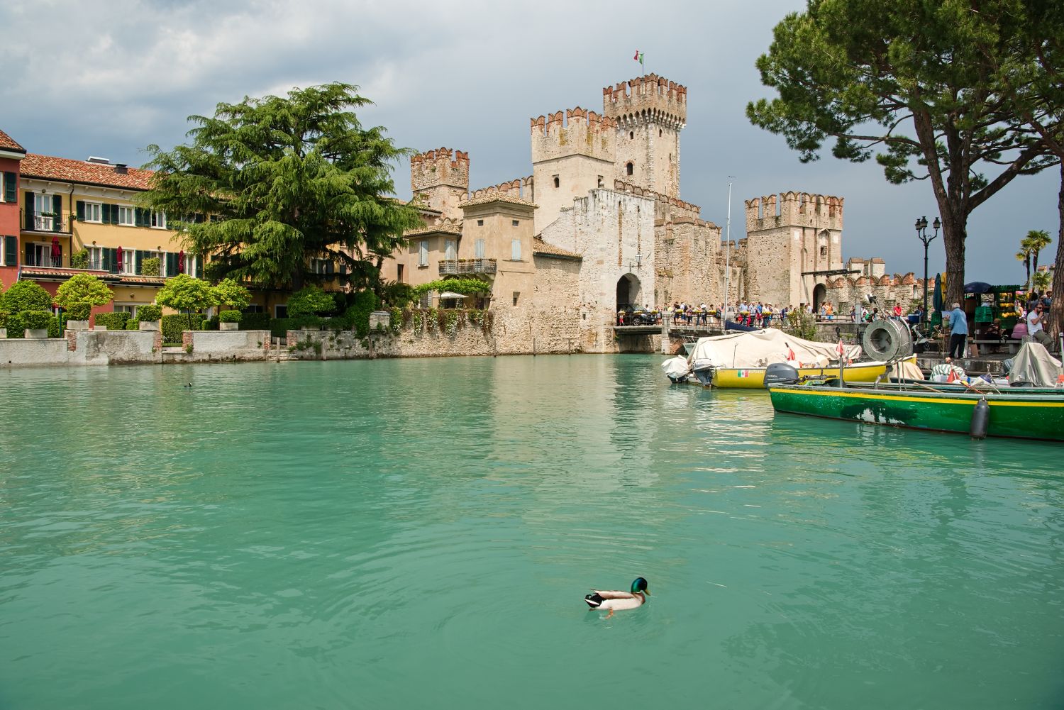 11 beautiful lakes in Northern Italy 6 A duck swims in teal water near colorful buildings and a historic stone castle with towers and battlements. Boats are docked nearby, and trees and people line the waterfront under a cloudy sky.