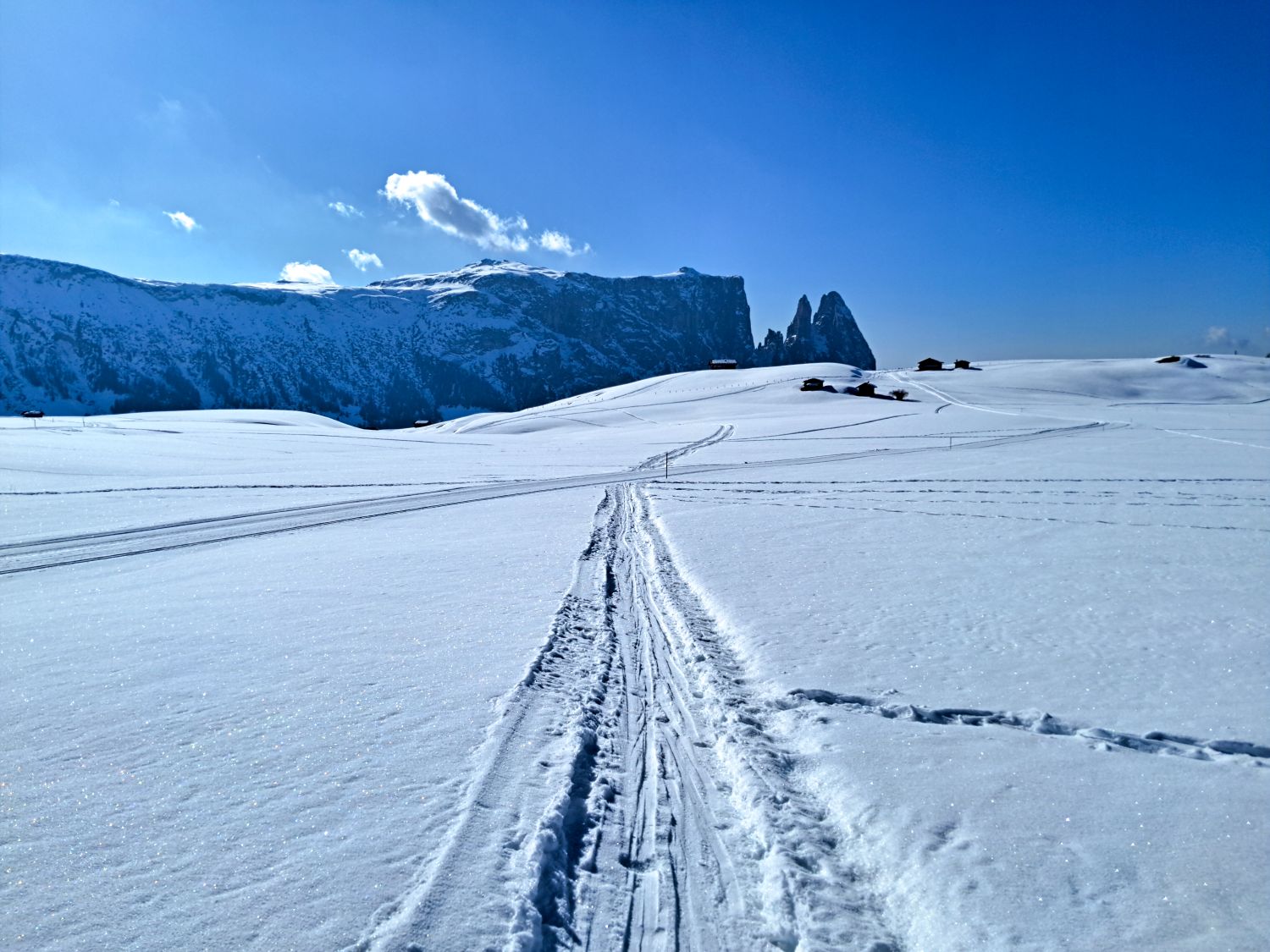 Dolomites in Winter: Weather, Skiing, Hiking, and more 12 Snow covered landscape with narrow ski tracks leading into the distance toward a range of rugged mountains under a bright blue sky. A few small wooden huts sit scattered on the rolling hills, emphasizing the quiet and remote winter setting.