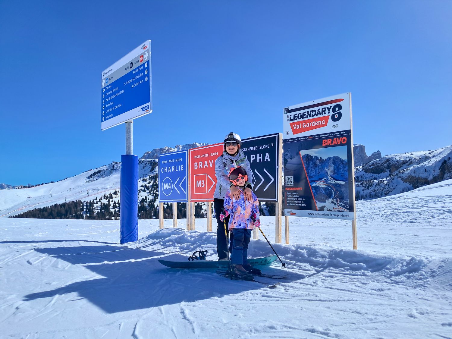 Where to stay in the Dolomites for skiing 3 Adult and child in ski gear stand on a snowy slope in front of trail signs while smiling at the camera. Signs behind them read "CHARLIE M4" "BRAVO M3" and "ALPHA" with directional arrows and a larger board that says "THE LEGENDARY 8 Val Gardena" and "BRAVO". Snow covered mountains and a clear blue sky stretch across the background.