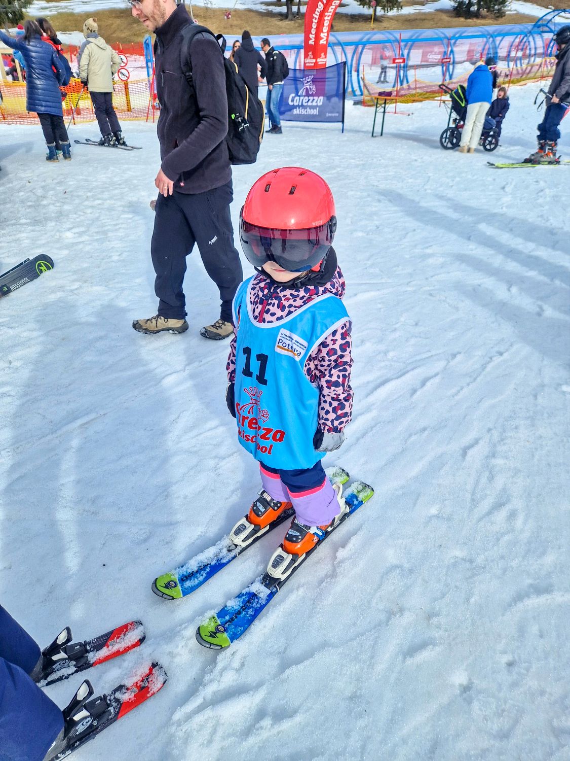 Skiing in the Dolomites for the first time 16 A young child in a red helmet, blue ski vest, and colorful jacket stands on skis in the snow, ready for a ski lesson. Adults and other skiers are nearby on a snowy slope with equipment and banners visible in the background.