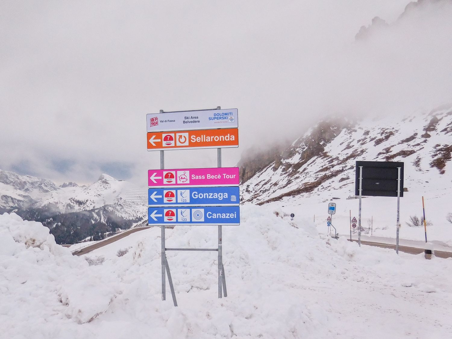 Skiing in the Dolomites for the first time 5 Directional signs for Sellaronda, Sass Becè Tour, Gonzaga, and Canazei stand in a snowy mountain landscape with cloudy skies and snow-covered peaks in the background.