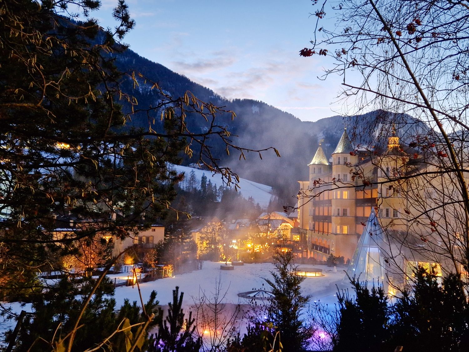 A warmly lit alpine hotel is nestled in a snowy valley at dusk, surrounded by dark evergreen trees and mountain silhouettes. Soft lights glow from the buildings and outdoor areas, with mist rising into the cold evening air, creating a cozy winter resort atmosphere.
