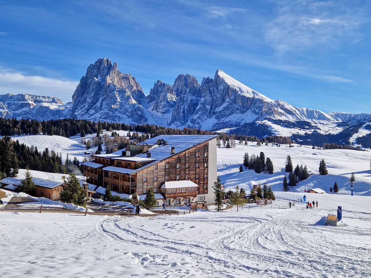 Dolomites in Winter: Weather, Skiing, Hiking, and more 28 A modern alpine lodge sits in a wide snowy meadow with ski tracks leading toward it, surrounded by scattered pine trees. Towering jagged mountain peaks rise sharply in the background under a bright blue winter sky, emphasizing the dramatic high alpine setting.