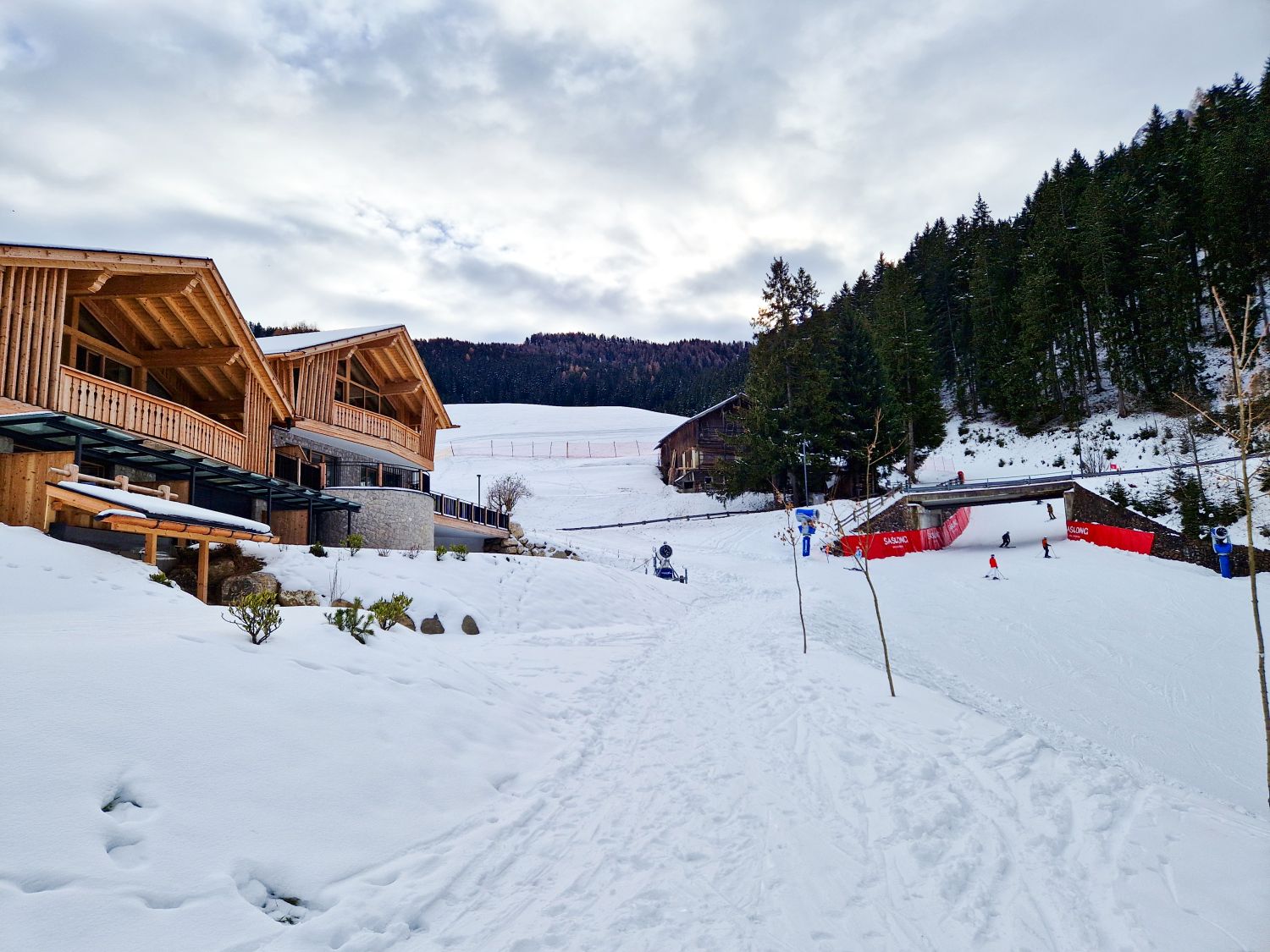 Where to stay in the Dolomites for skiing 11 A snowy ski resort with wooden lodges on the left, people skiing on a slope to the right, and pine trees lining the hillside. The sky is cloudy and the area is surrounded by snow-covered ground.