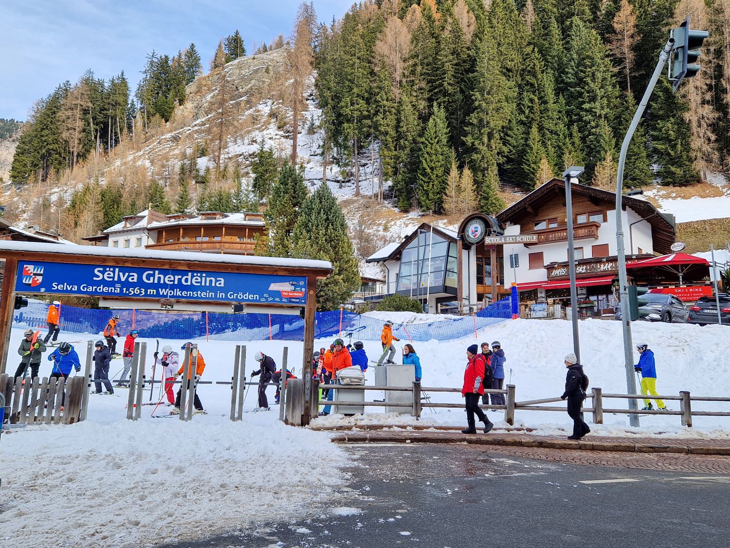 Skiing in the Dolomites for the first time 7 A busy ski school area in Selva Gardena shows skiers lining up beneath a large sign reading Selva Gherdeina Selva Gardena 1,563 m Wolkenstein in Gröden. People in colorful ski gear gather near beginner slopes and fencing, with alpine buildings and forested mountains rising behind the village setting.