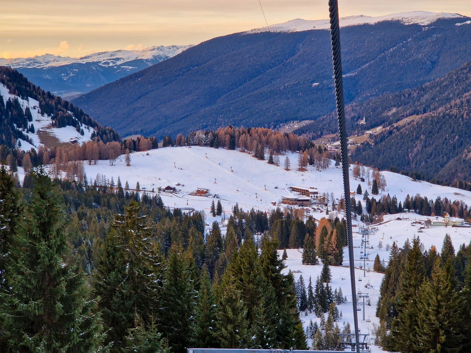 Things To Do In The Dolomites In Winter: Ski And No Ski 9 A ski area sits in a snowy valley with chairlift lines descending through evergreen trees toward lodges and open beginner slopes. Forested mountains rise in layered ridges beyond the resort, with warm late day light touching the distant peaks.