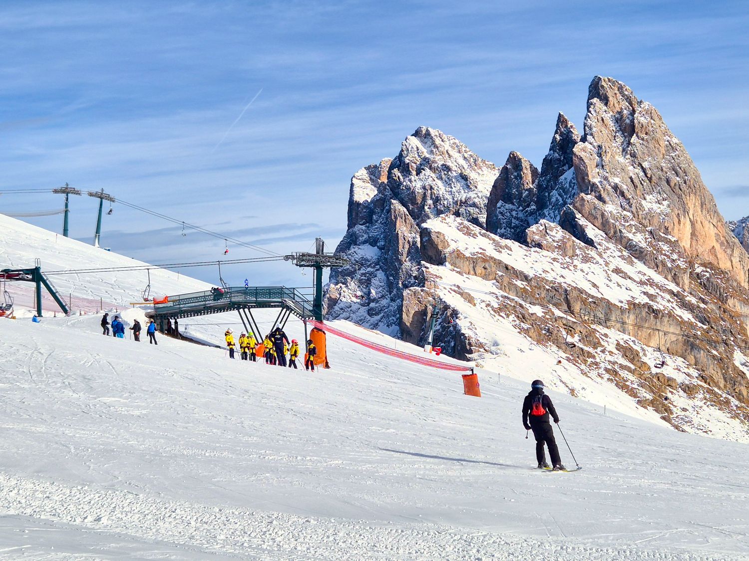 Where to stay in the Dolomites for skiing 12 A ski lift station sits on a snowy slope with a small group of skiers gathered nearby while one skier glides downhill in the foreground. Jagged rocky mountain peaks rise sharply behind the lift under a clear blue winter sky, emphasizing the dramatic high alpine setting.