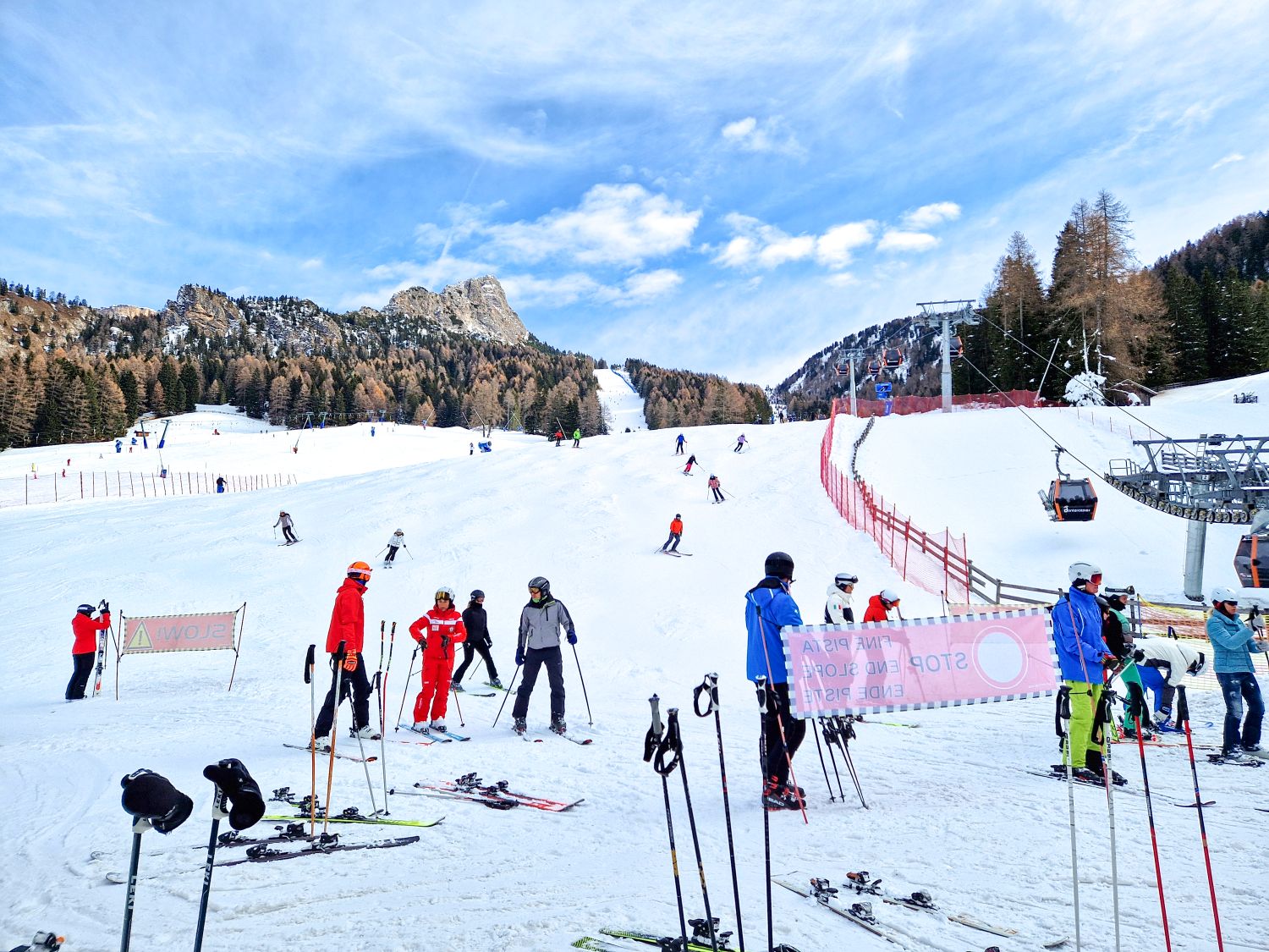 Where to stay in the Dolomites for skiing 15 A wide ski slope is busy with skiers gathered at the bottom while others descend from above under a bright blue sky. Skis and poles rest in the snow near warning signs, with chairlifts, safety fencing, and forested mountains framing an active winter resort scene.