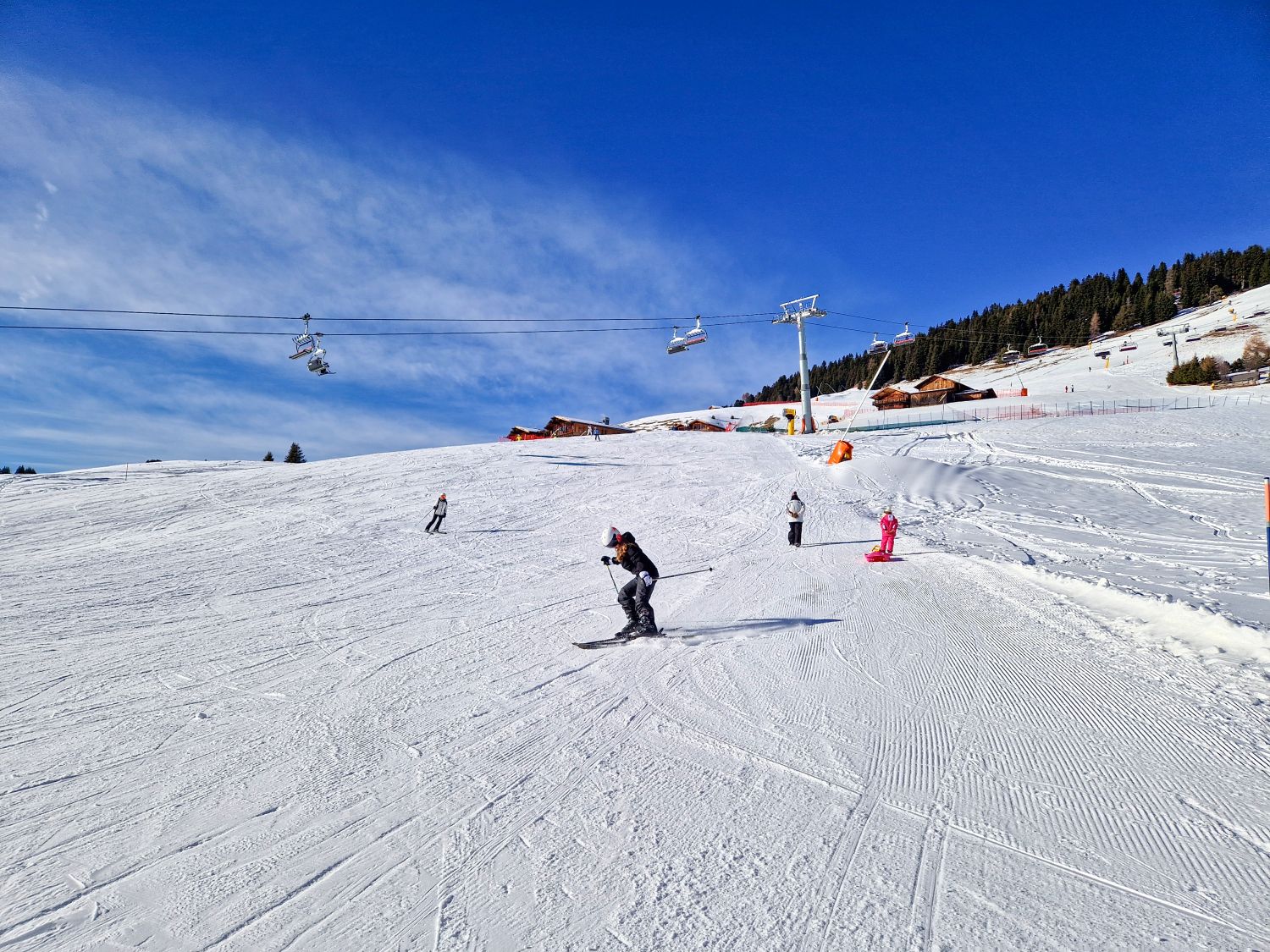 Where to stay in the Dolomites for skiing 4 A groomed ski slope slopes gently downhill with several skiers and a snowboarder moving across the snow. Chairlifts pass overhead toward small alpine huts at the top of the run under a deep blue winter sky.