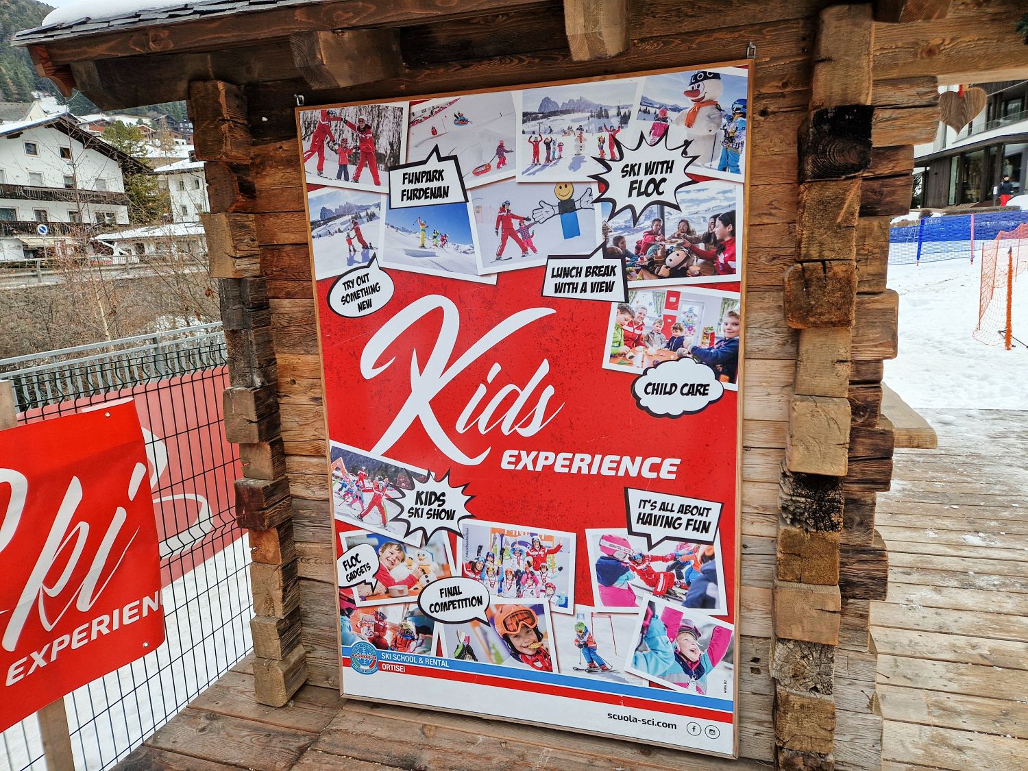 Skiing in the Dolomites for the first time 17 A large red poster titled Kids Experience is mounted on a rustic wooden structure at a ski school area. The poster includes photos and text reading Funpark Furdenan, Ski with Floc, Lunch break with a view, Child care, Kids ski show, Final competition, Try out something new, and It’s all about having fun.