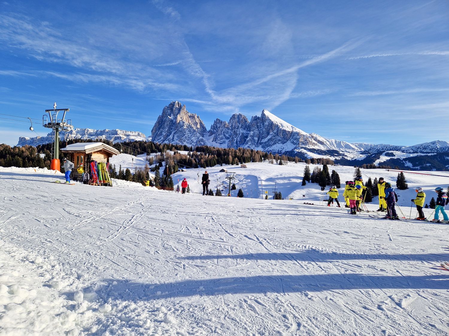 Skiing in the Dolomites for the first time 8 A wide beginner ski area opens onto a panoramic alpine landscape, with children in bright ski gear gathered for a lesson on a gentle slope. Snow covered meadows and evergreen trees stretch toward dramatic jagged mountain peaks under a vivid blue winter sky.