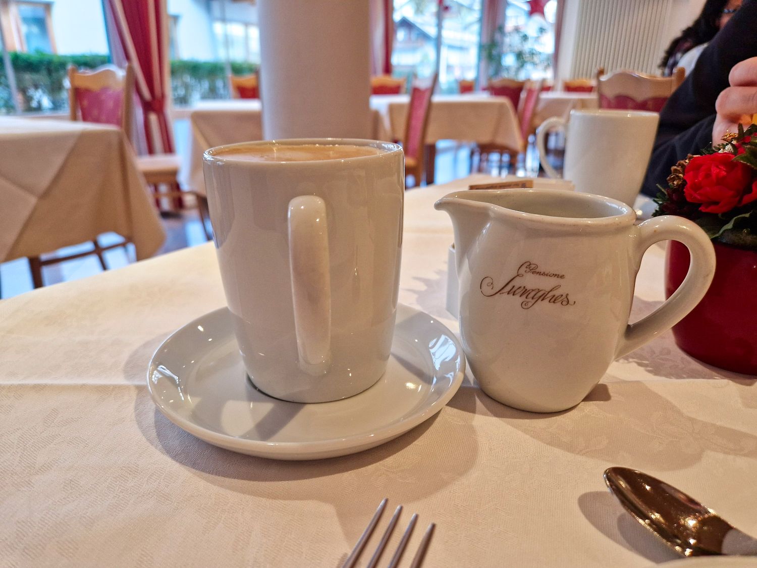 A cup of coffee on a saucer sits next to a small white creamer jug on a table in a cozy café, with tables, chairs, and a window with red curtains in the background.