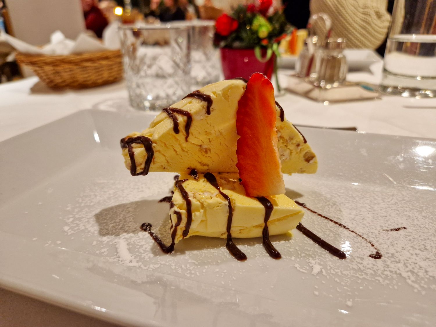 A dessert featuring two slices of yellow ice cream drizzled with chocolate sauce, garnished with a strawberry slice, and dusted with powdered sugar, served on a white plate in a restaurant setting.