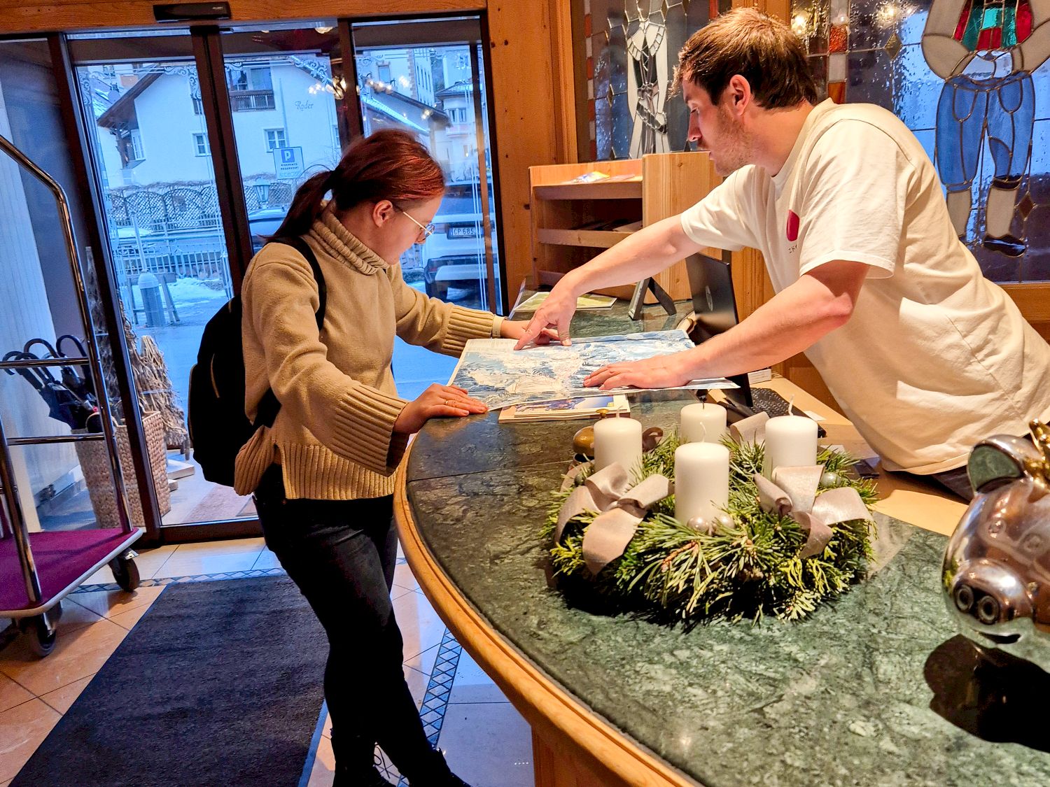 A hotel receptionist points to a map on the counter while assisting a woman wearing glasses and a tan sweater. A decorative candle wreath sits on the marble counter near them. Large windows are in the background.