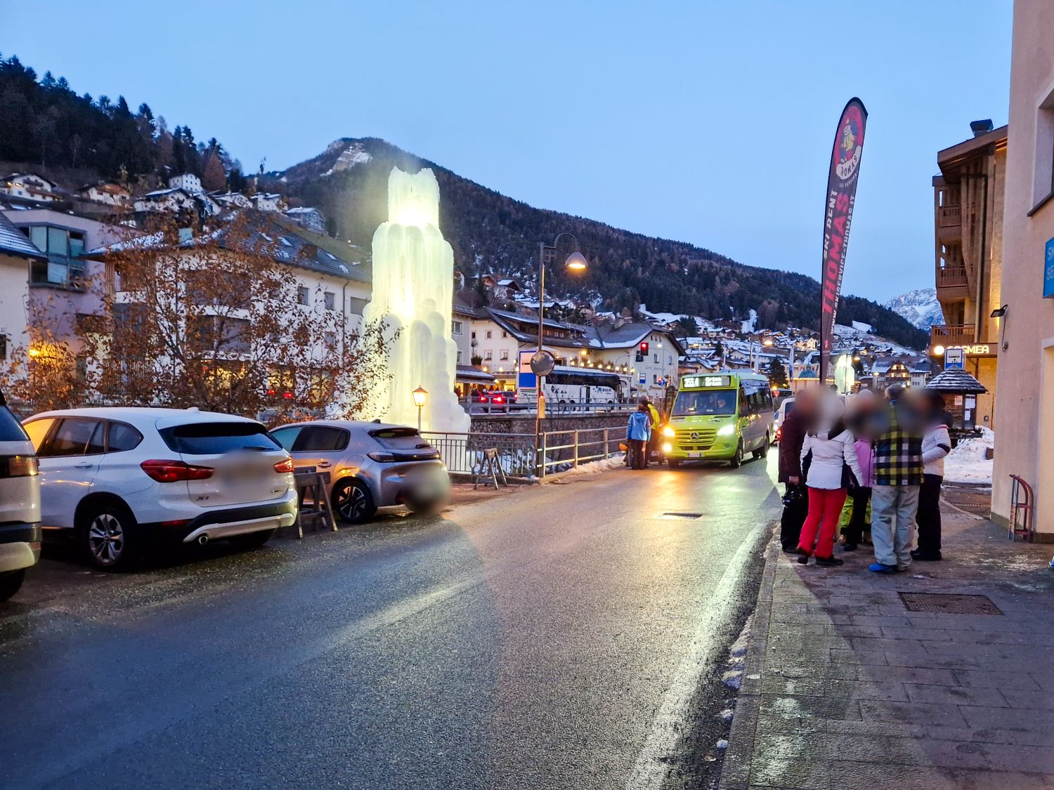 A group of people waits near a bus stop on a winter street lined with parked cars, buildings, and a glowing ice sculpture; snow covers the surrounding area and mountains are visible in the background.
