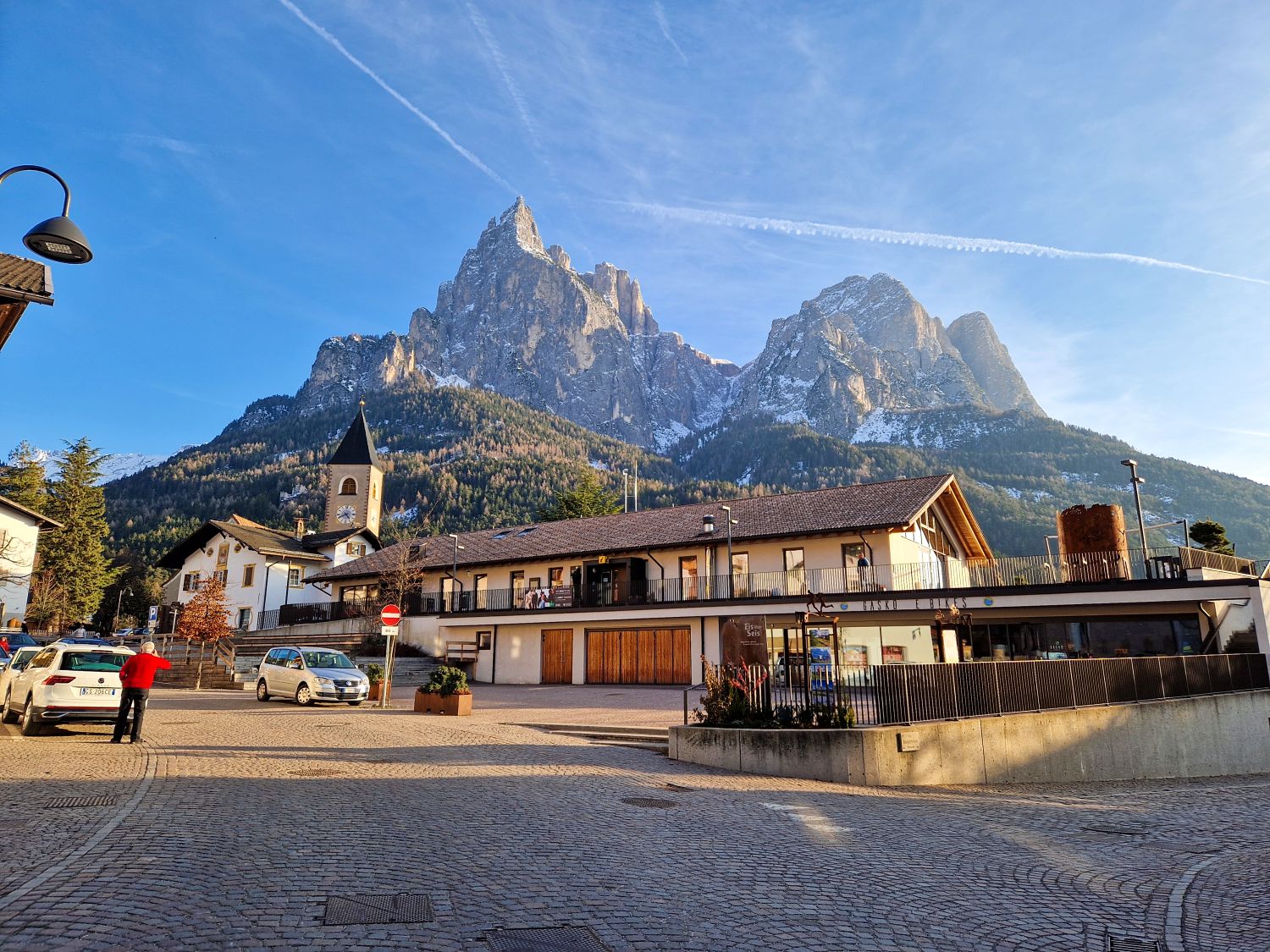 Where to Stay in the Dolomites in The off-season 6 A quiet village square with a church tower and low buildings sits beneath dramatic jagged mountain peaks dusted with snow under a bright blue sky. A person in a red jacket stands near parked cars, adding scale to the towering alpine landscape behind the town.