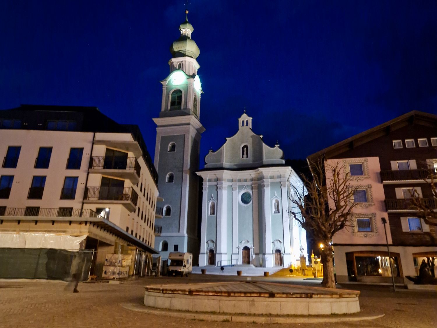 Where to Stay in the Dolomites in The off-season 4 Nighttime view of a church with a tall bell tower and onion dome illuminated against a deep blue sky in a quiet town square. The pale facade of the church stands between modern alpine buildings with bare trees and empty pavement, creating a calm evening atmosphere.