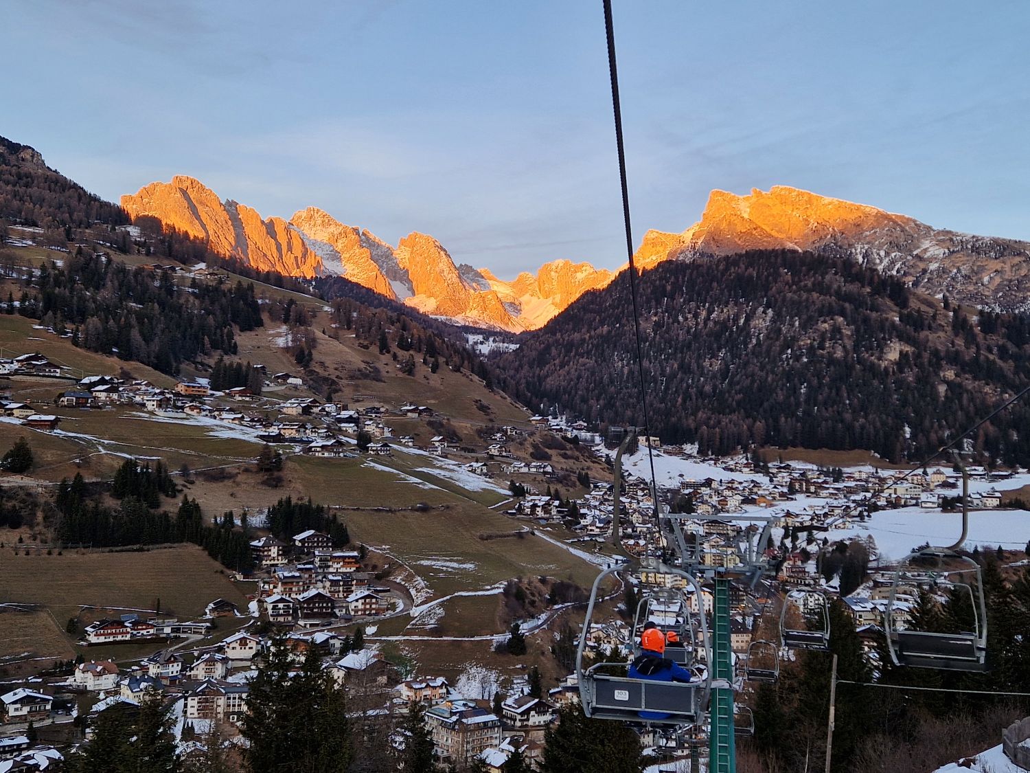 Dolomites in Winter: Weather, Skiing, Hiking, and more 19 Winter view in Santa Cristina showing a chairlift ascending over snowy slopes with glowing pink alpenglow on the Dolomite peaks during enrosadira. Soft pastel light reflects off the mountains at sunset, creating a calm alpine winter atmosphere.