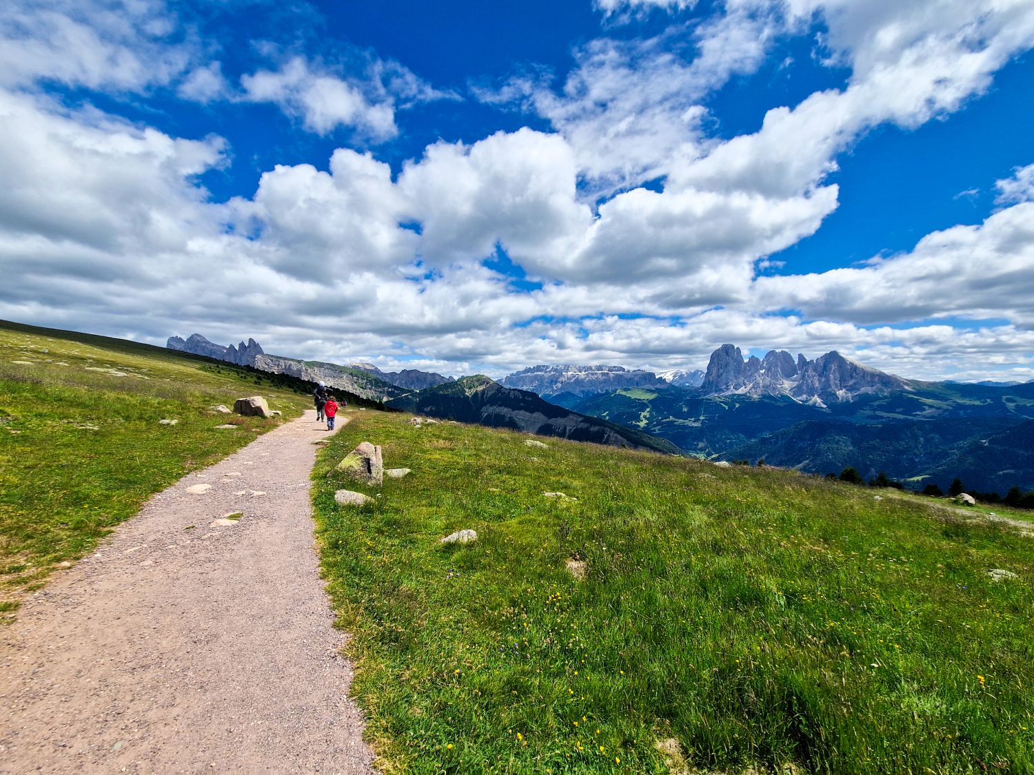 Best Dolomites Hiking Shoes: Boots, Trail runners, other? 3 A wide gravel hiking path winds through a green alpine meadow with two hikers walking toward distant jagged mountain peaks. Rolling hills, rocky outcrops, and dramatic cliffs stretch across the horizon under a bright blue sky filled with large white clouds. The image conveys an open scenic mountain landscape and a peaceful hiking experience in nature.