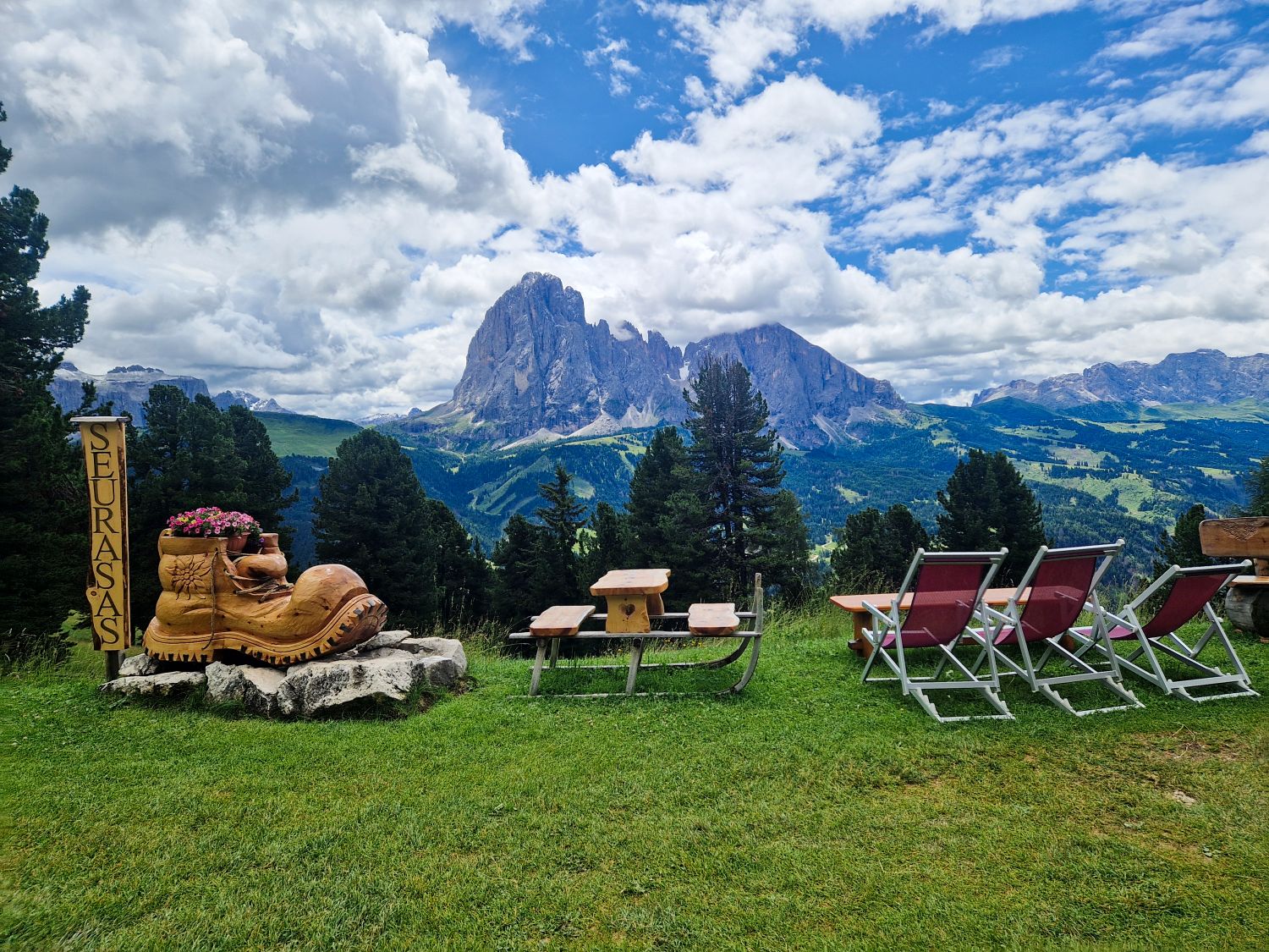 Summer view near Santa Cristina in the Dolomites with a traditional alpine hut at Baita Seurasas surrounded by green meadows and rolling hills. Forested slopes and rugged mountain peaks rise in the background under a clear July sky, showing a peaceful mountain hiking landscape.