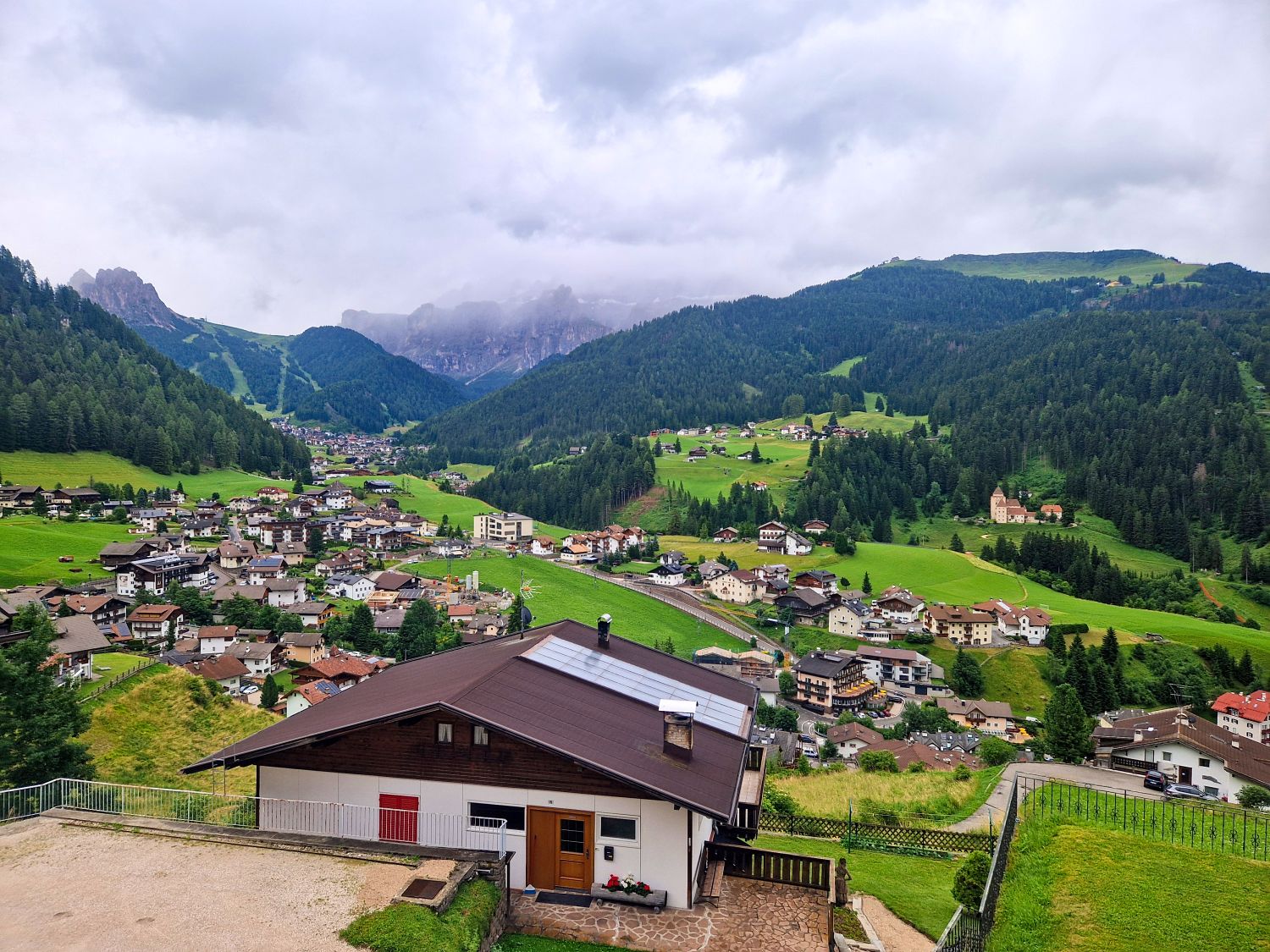 Summer mountain view near Santa Cristina with the Sella Group rising behind green alpine meadows around Baita Seurasas. Rolling hills, scattered trees, and dramatic rocky peaks create a wide open Dolomites landscape on a clear July day.