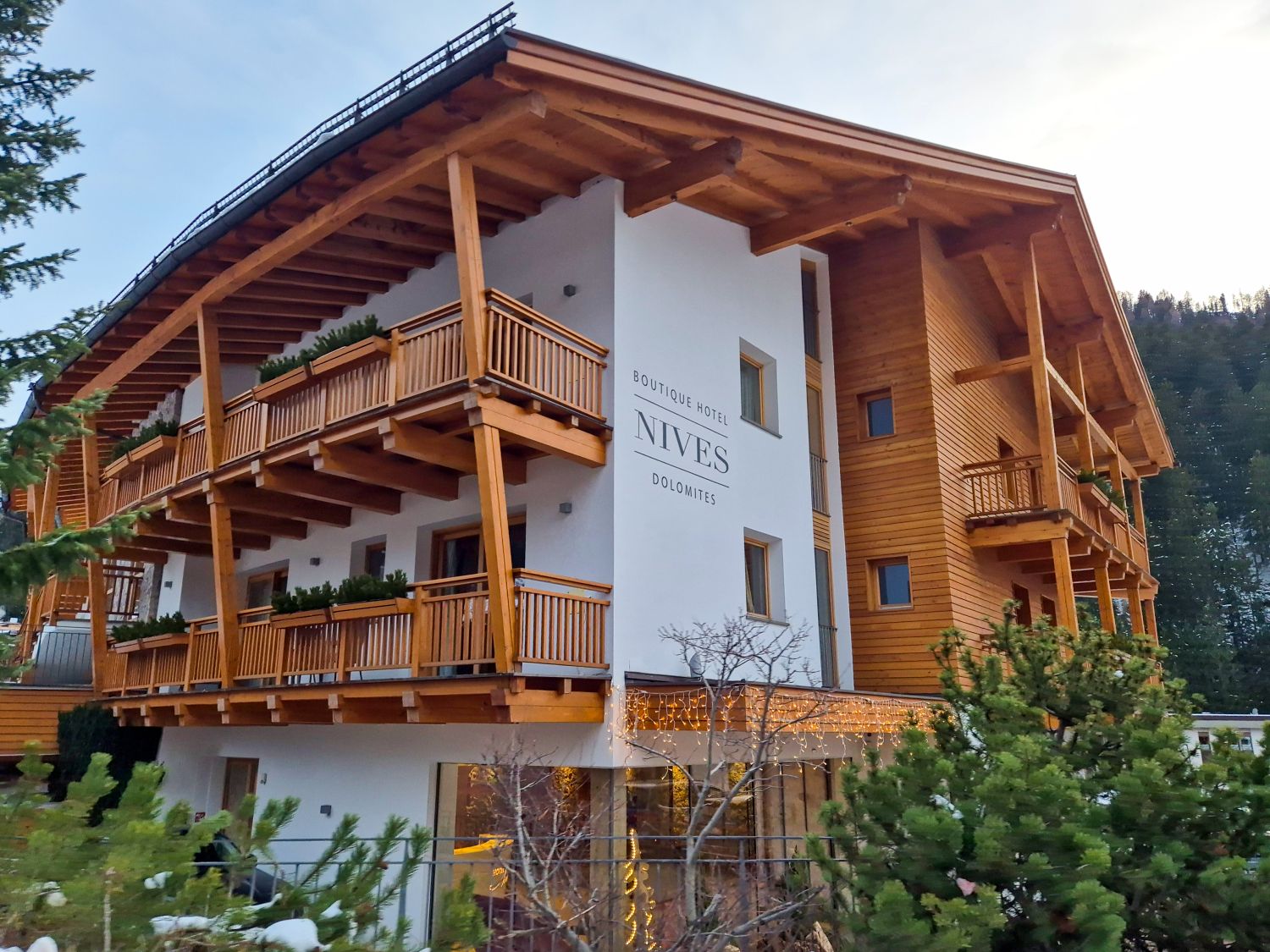 Modern alpine hotel with wooden balconies and sloped roof stands against a forested mountain backdrop. The white exterior wall displays the text "BOUTIQUE HOTEL NIVES DOLOMITES" and warm lights glow from the ground floor windows.