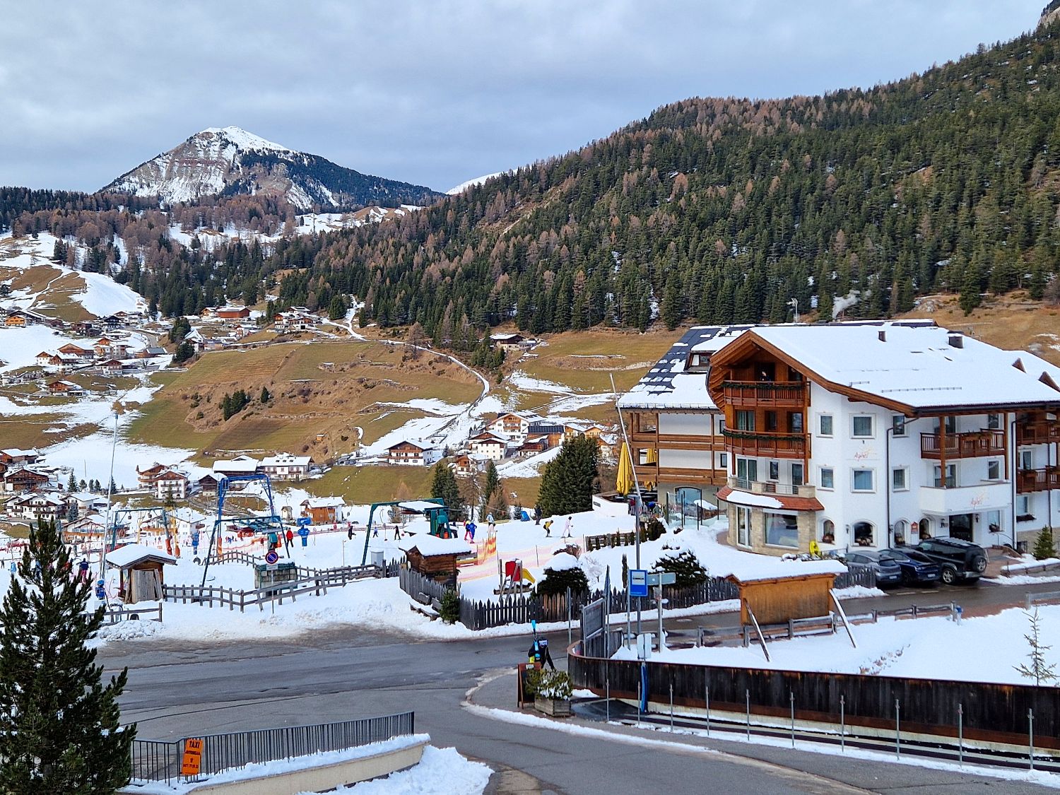 Snow covered alpine village Selva di Val Gardena spreads across a valley with scattered wooden houses and winding roads between hills. In the foreground a hotel sits beside a small ski area where people gather near lifts, with forested mountains and a partially snow capped peak in the background.