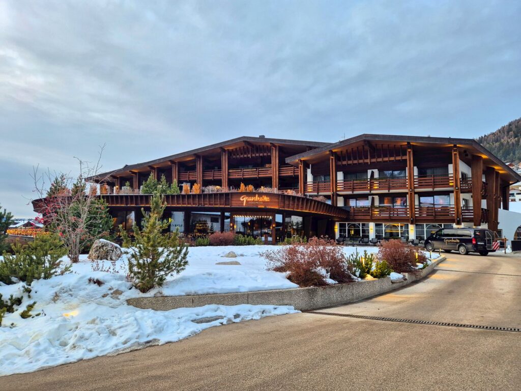 Modern alpine hotel with wooden balconies and warm lighting at the entrance, with a sign reading "Granbaita Dolomites" above the doorway. A curved driveway leads past snow covered landscaping and shrubs toward the building under an overcast evening sky.