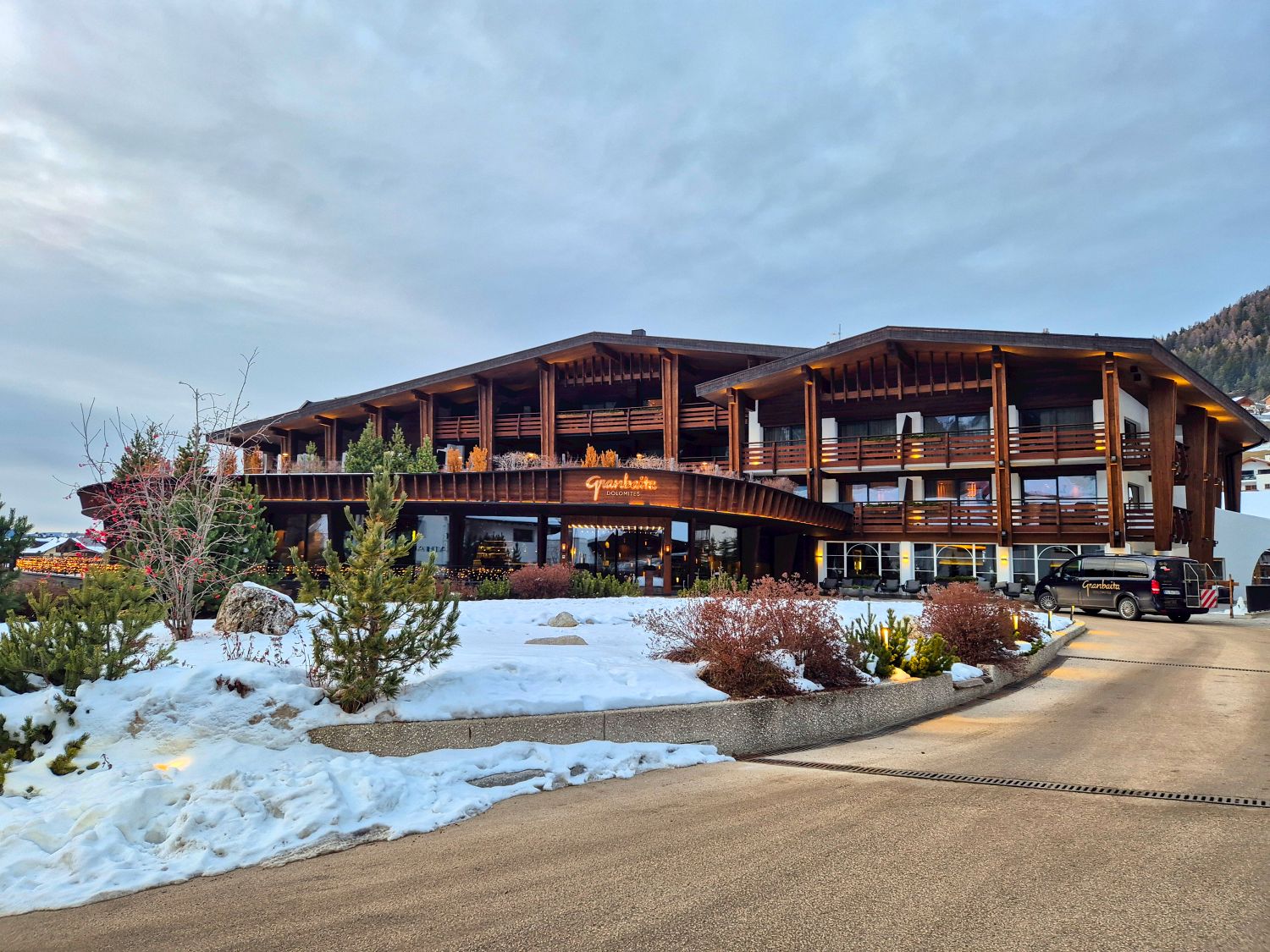 Modern alpine hotel with wooden balconies and warm lighting at the entrance, with a sign reading "Granbaita Dolomites" above the doorway. A curved driveway leads past snow covered landscaping and shrubs toward the building under an overcast evening sky.