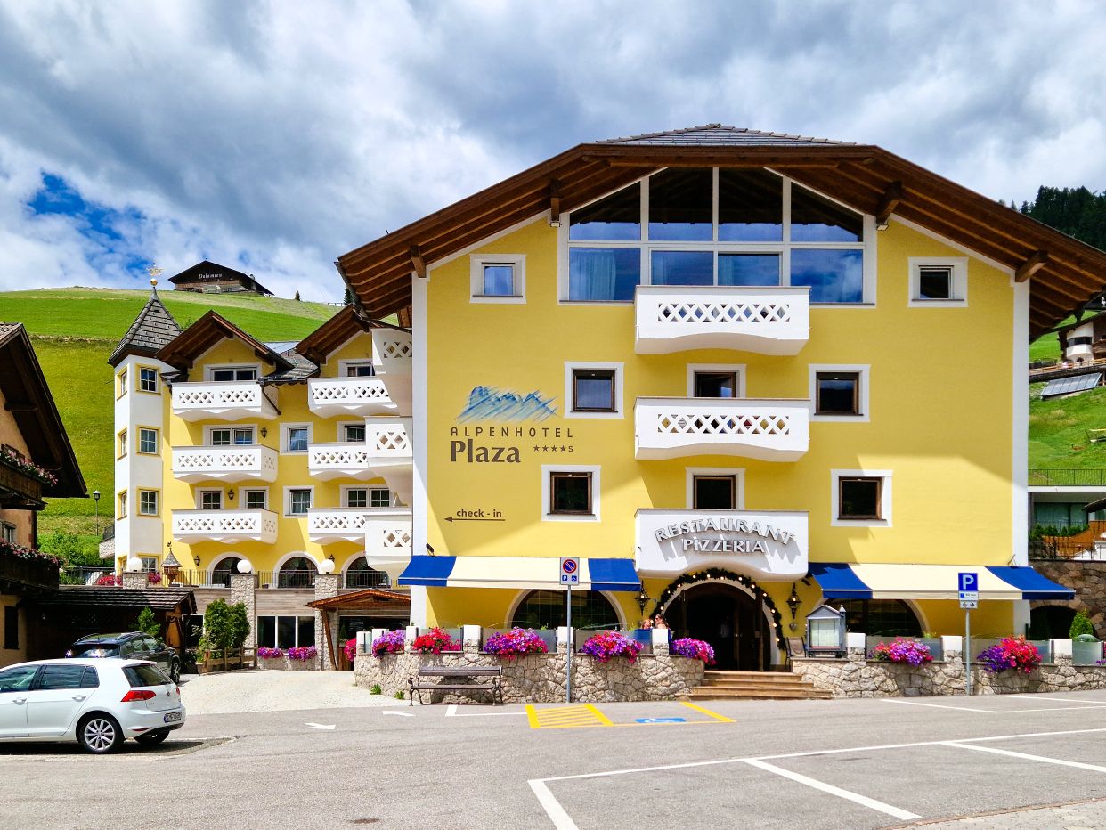 Front view of Alpenhotel Plaza in Val Gardena featuring white balconies, blue awnings, vibrant flower boxes, and the entrance to the restaurant and pizzeria. Traditional alpine homes and rolling green hills frame the hotel beneath a cloudy mountain sky, highlighting the charm of hotels in Val Gardena.