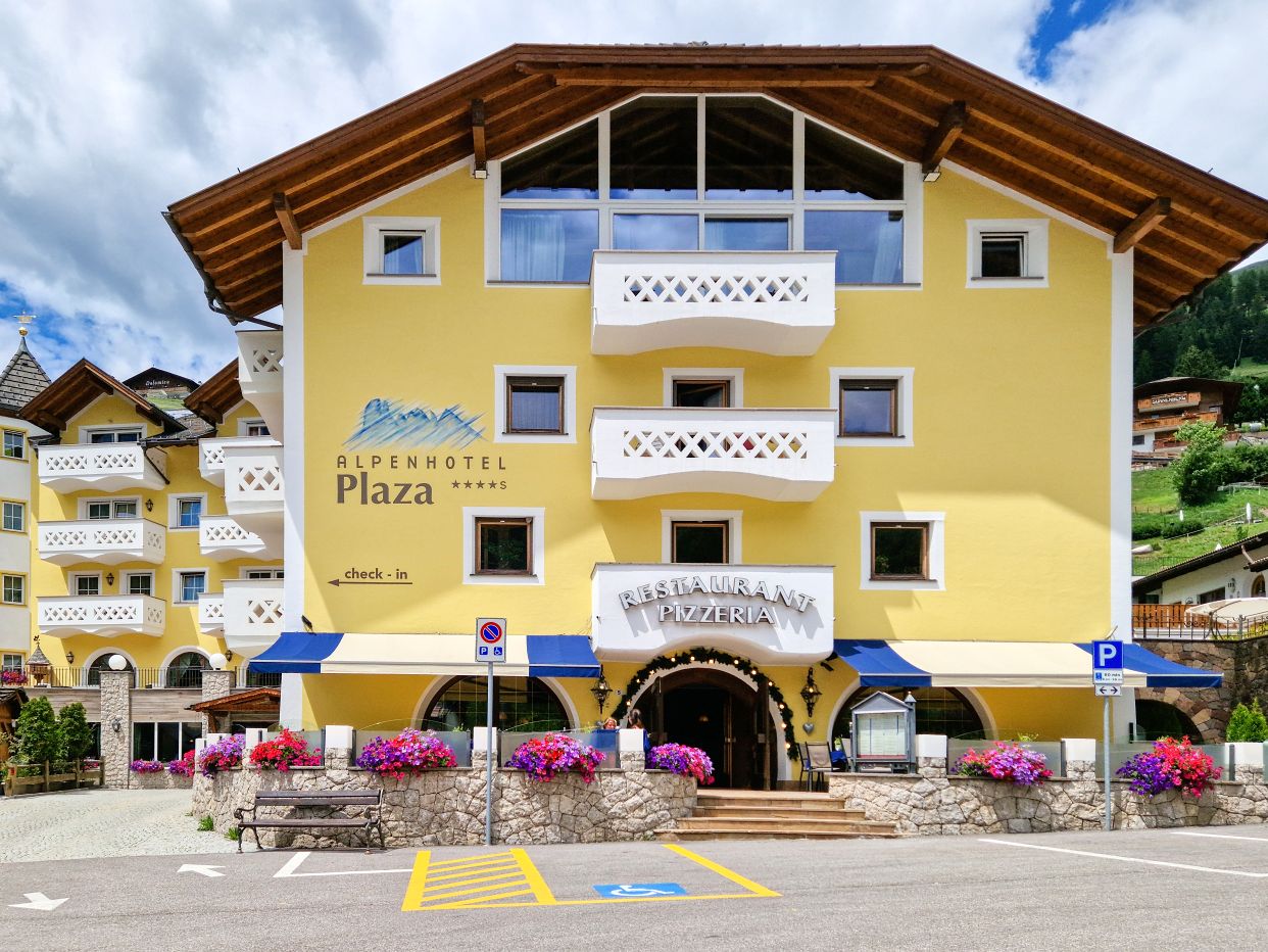 Alpenhotel Plaza in Val Gardena with pale yellow walls, white balconies, and a restaurant entrance lined with colorful flower boxes, viewed from across a parking area. Green alpine hills and traditional mountain buildings surround the hotel under a dramatic cloudy sky, showcasing one of the hotels in Val Gardena.