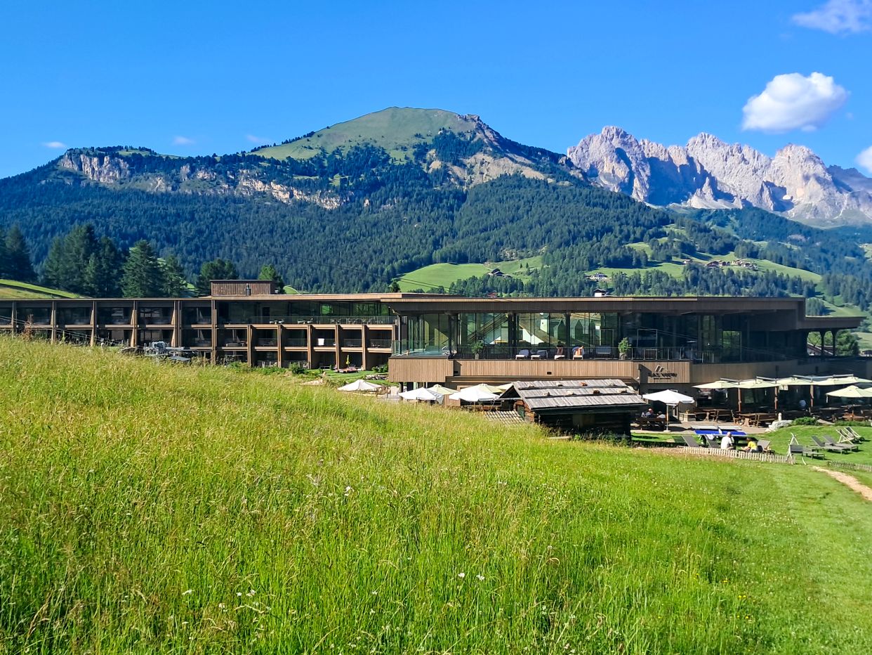 Hotel Cendevaves in Santa Cristina sits at the edge of a green alpine meadow with a modern wood and glass exterior and outdoor terraces. The dramatic peaks of the Dolomites rise all around the property beneath a bright blue sky, highlighting one of the scenic hotels in Val Gardena.