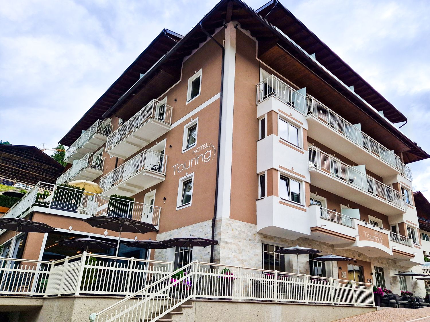 Hotel Touring in Santa Cristina features a modern alpine design with warm brown walls, white balconies, and a spacious outdoor terrace with umbrellas. Surrounded by mountain scenery, it is one of the welcoming hotels in Val Gardena, offering contemporary accommodations in the heart of the village.