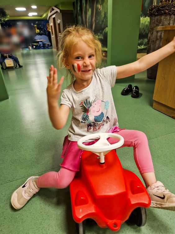 Emma (young child) sits on a small red ride on toy indoors, smiling and raising one hand while showing face paint in green white and red on their cheek. The play area has green floors and walls with other children and toys visible in the background.
