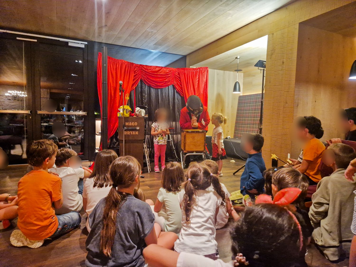 Children sit on the floor watching a small indoor magic show as a performer in a red jacket leans over a wooden box on stage. Red curtains frame the setup and a sign reads "MAGO BRYAN" while two children assist during the performance. Magic show at Hotel Biancaneve - one of the best family hotels in Val Gardena