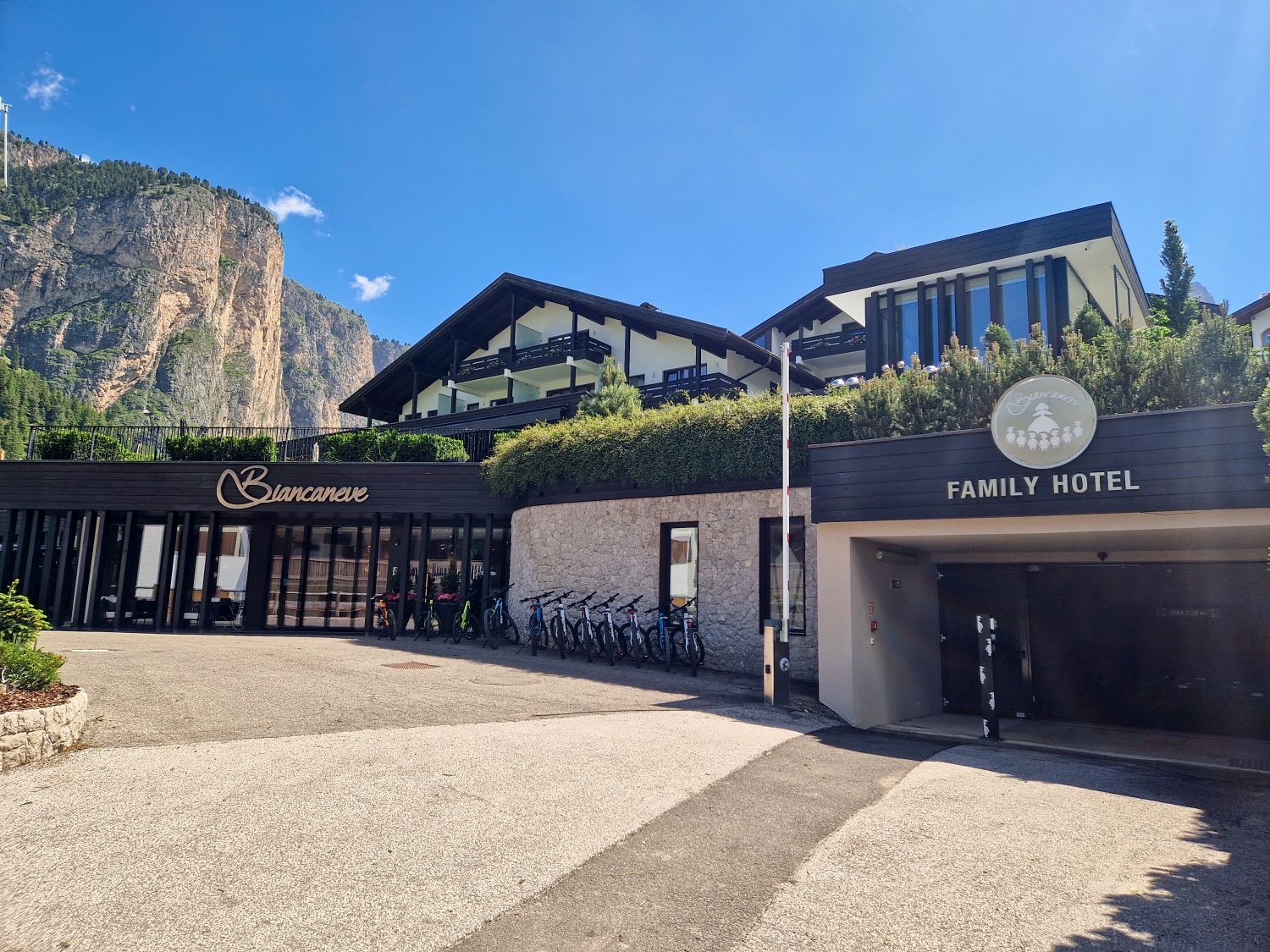 Contemporary alpine hotel entrance with dark wood facade and glass doors sits beneath a cliffside backdrop and clear blue sky. Signage reads "Biancaneve" and "FAMILY HOTEL" with a row of bicycles parked outside along the stone wall.
