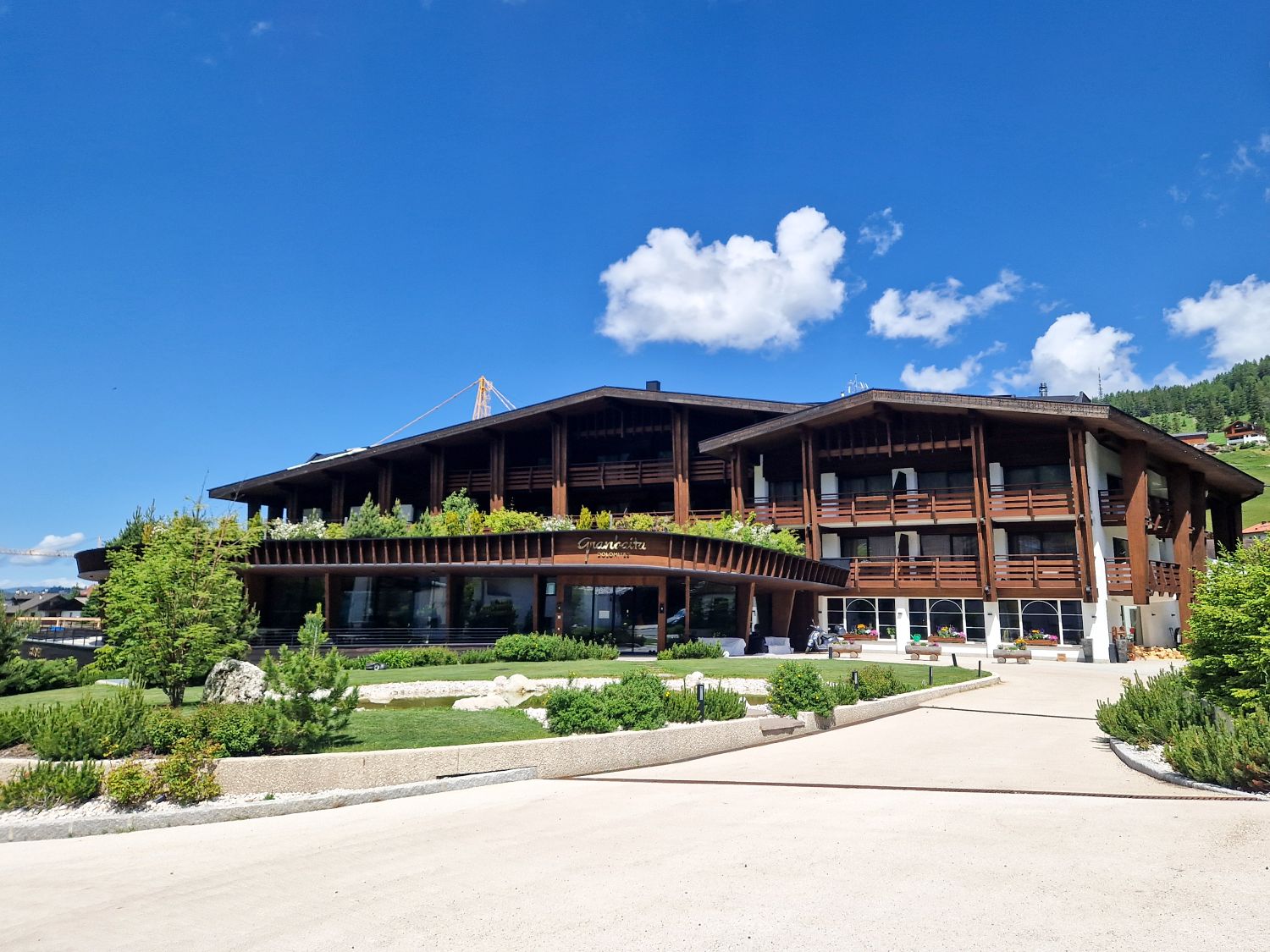 Large modern alpine hotel with dark wood beams and wide balconies faces a landscaped garden and curved driveway under a bright blue sky. The entrance is framed by greenery and glass windows, with the name "Granvara" displayed above the main doors.