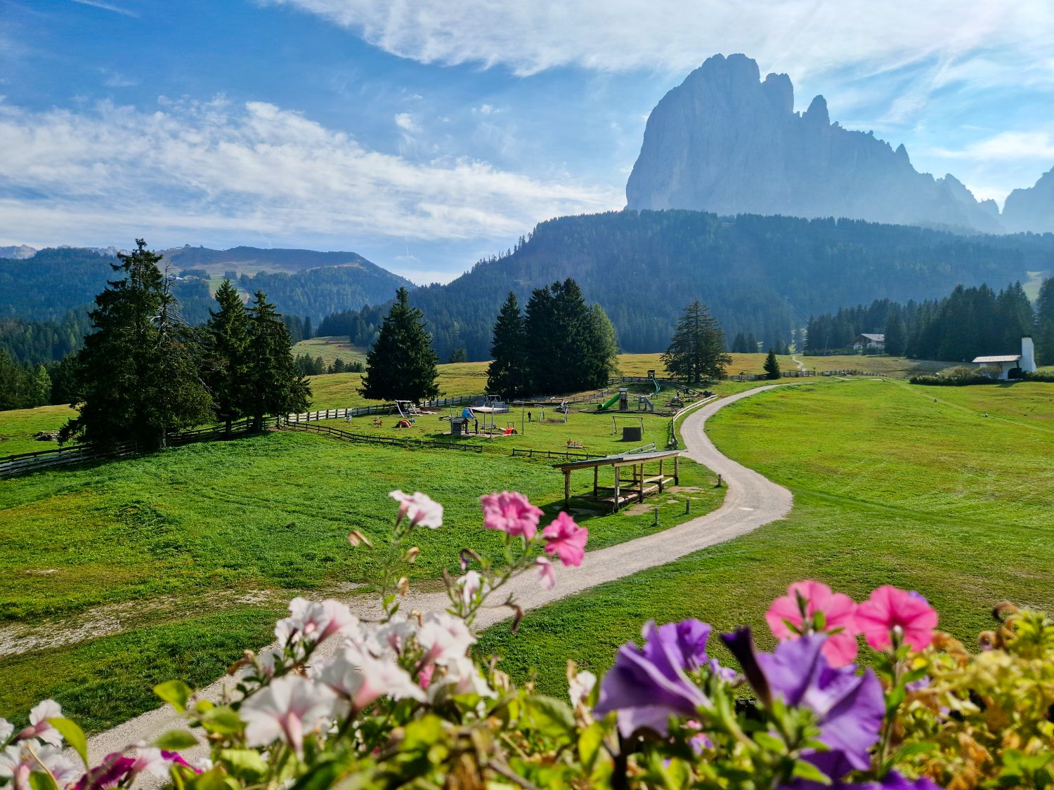 Where to Stay in the Dolomites in The off-season 7 Green meadow in front of hotel Monte Pana with a winding path leading past a small playground and wooden shelters toward distant alpine mountains. Colorful flowers in the foreground frame the view with tall trees and a dramatic rocky peak rising in the background.