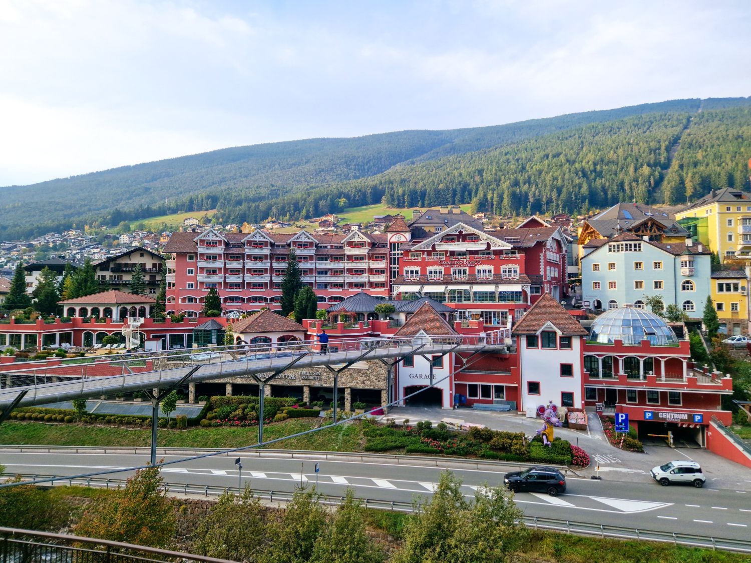 Large red and white alpine resort complex with balconies and decorative trim set against a green hillside. A pedestrian bridge crosses over a road in front, and signs reading "GARAGE" and "CENTRUM PARKING" are visible near the entrance.