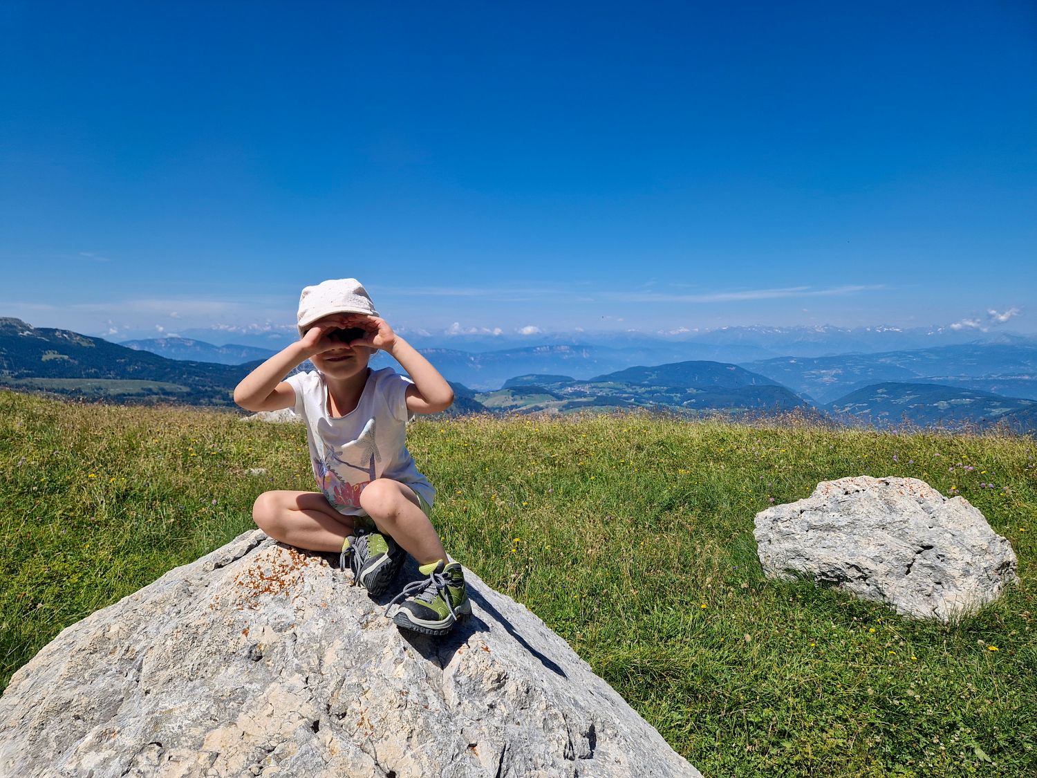 Best Dolomites Hiking Shoes: Boots, Trail runners, other? 9 Emma sits on a large rock along the Latemar Panorama Trail near Obereggen, shading her eyes as she looks out across rolling alpine meadows and distant mountain peaks. The clear blue sky and wide-open views capture the peaceful scenery of this family-friendly hike in the Dolomites.