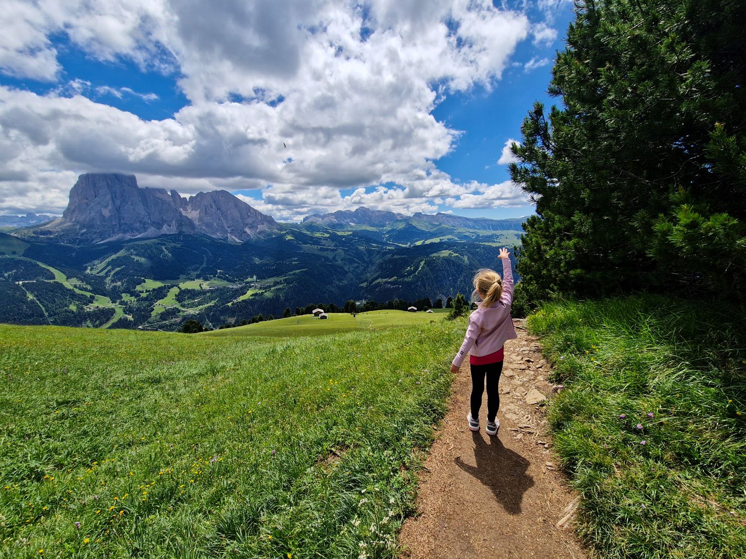 Best Dolomites Hiking Shoes: Boots, Trail runners, other? 2 Emma hikes toward Mt. Pic above Santa Cristina in Val Gardena wearing hiking sandals on a narrow trail through a green alpine meadow. The rolling hills and rugged Dolomite peaks illustrate the varied terrain where footwear with good sole grip, including sturdy hiking sandals, can be suitable for many routes in the Dolomites.