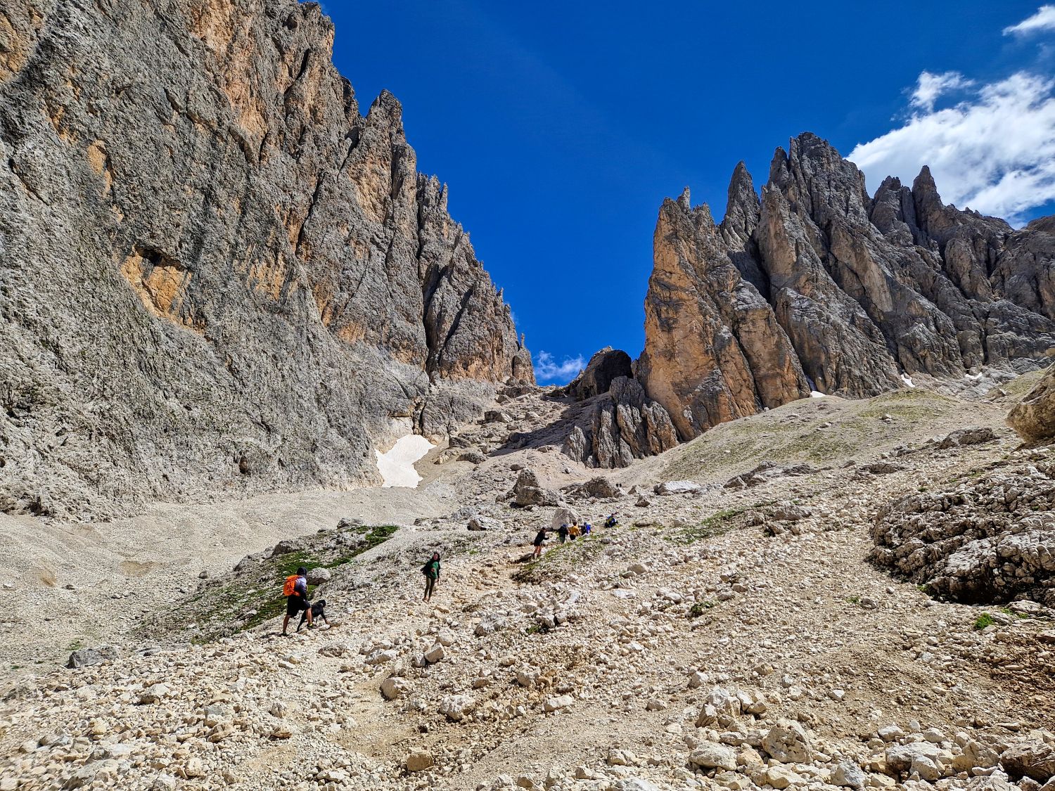 Best Dolomites Hiking Shoes: Boots, Trail runners, other? 6 Hikers make their way across the rocky scree slopes of the Sassolungo Loop hike in Val Gardena, surrounded by towering limestone cliffs and jagged Dolomite peaks. The rugged alpine landscape stretches beneath a deep blue sky, showcasing one of the region's most dramatic hiking routes.