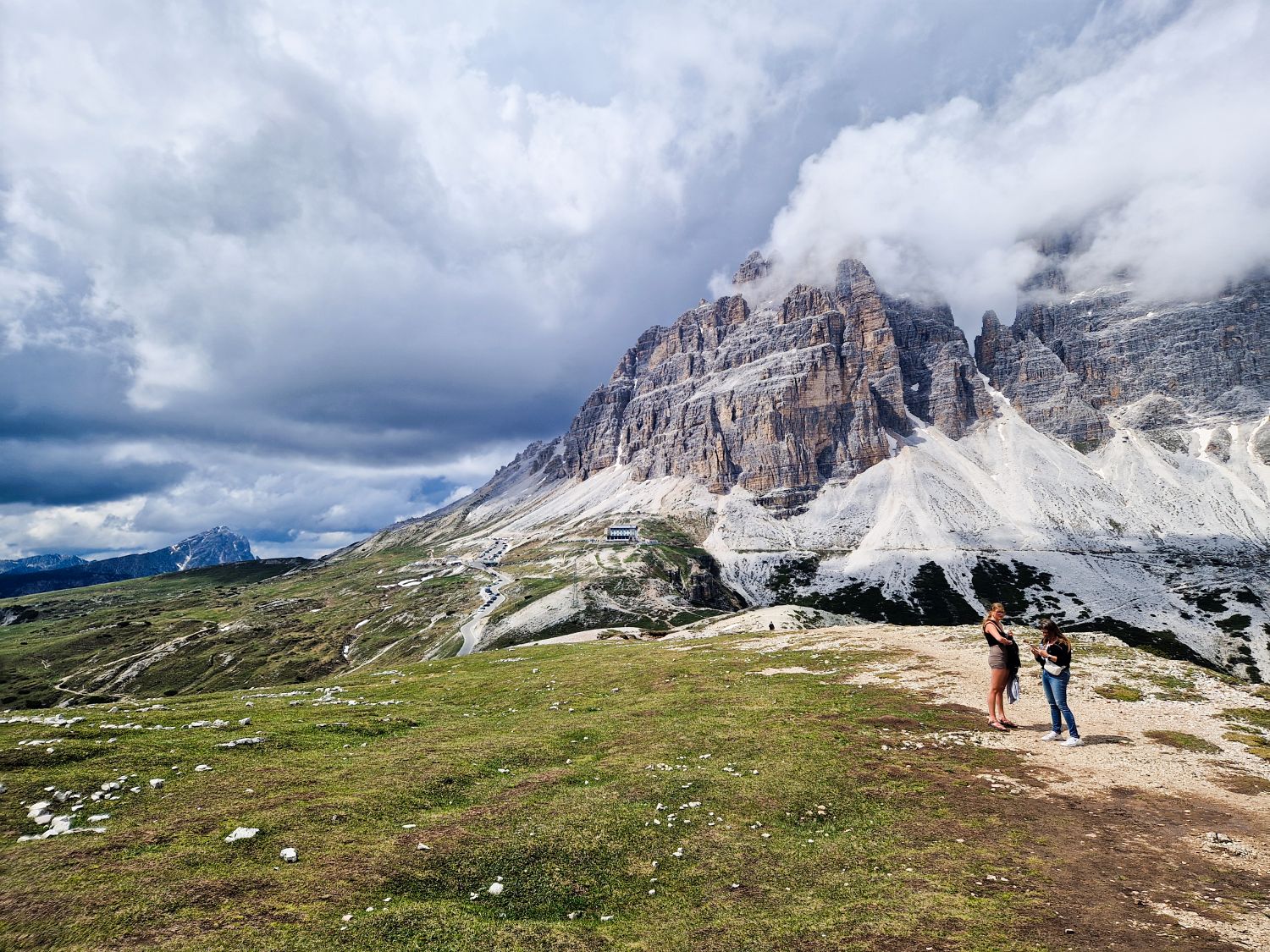 Best Dolomites Hiking Shoes: Boots, Trail runners, other? 8 Two visitors stand on the Cadini di Misurina viewpoint trail overlooking dramatic limestone peaks, with Tre Cime di Lavaredo partially hidden by low clouds in the background. Rifugio Auronzo is visible below among the rocky slopes, while dark storm clouds gather over the surrounding Dolomite landscape.