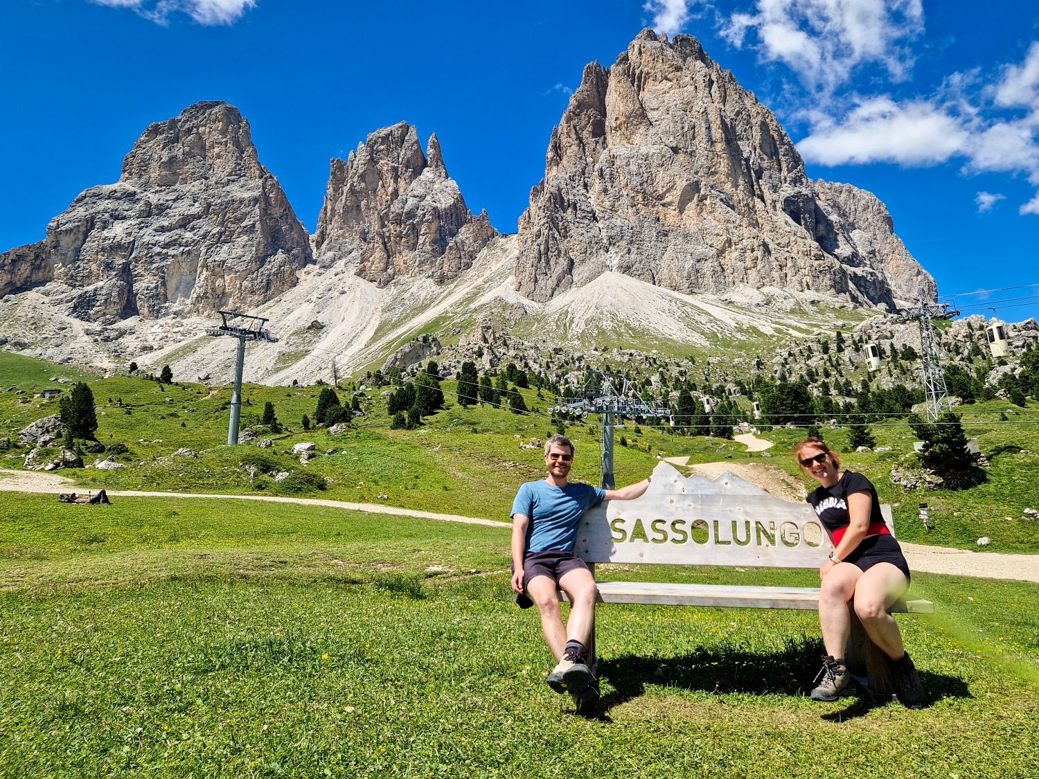 Kristine and Michal sit on a wooden bench with a sign reading “SASSOLUNGO” in front of the Sassolungo Group at Passo Sella in the Dolomites. Towering limestone peaks rise behind green alpine meadows, hiking paths, and cable car lines under a bright blue sky with scattered clouds.
